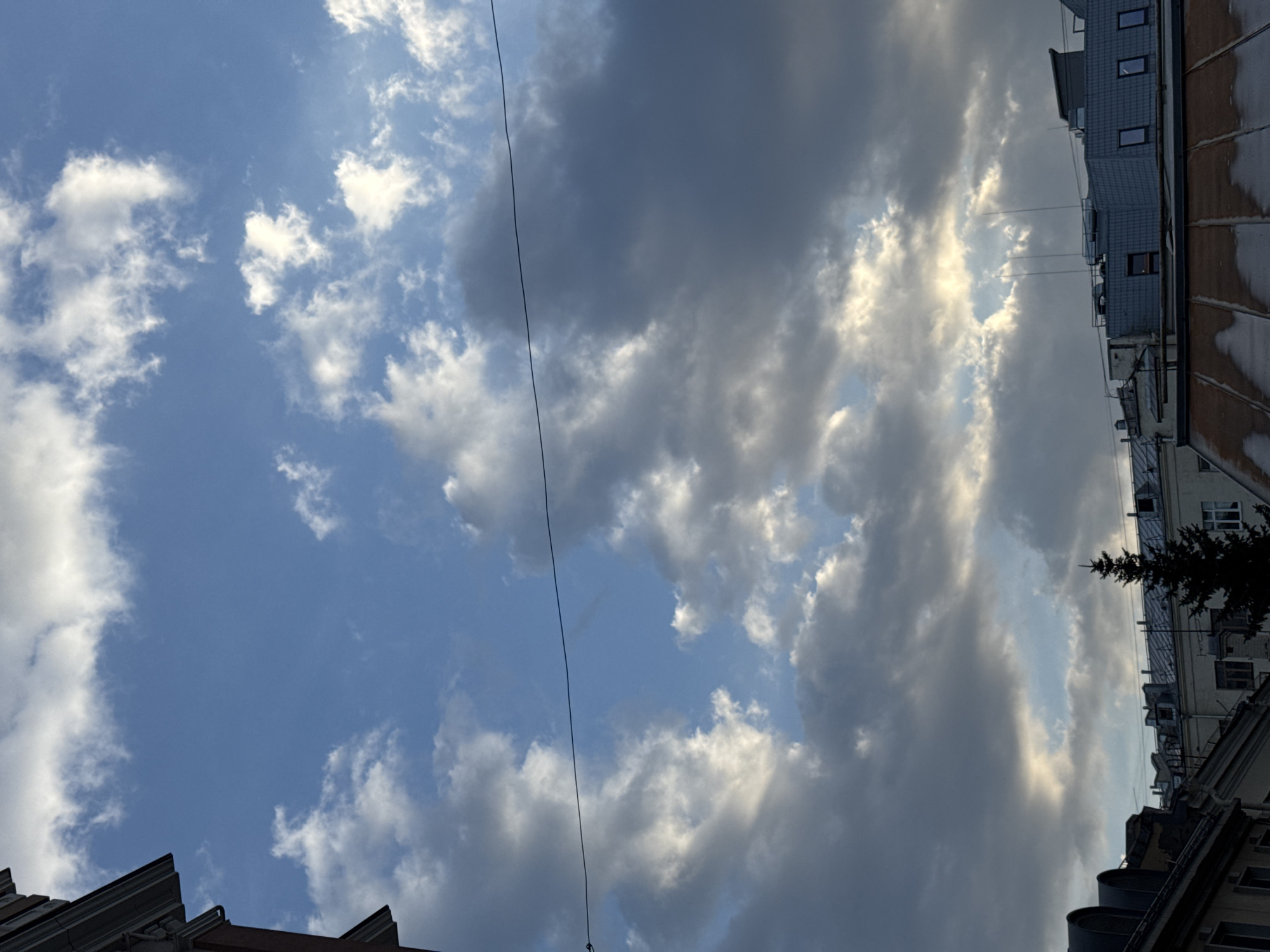 The image depicts a view of the sky above Moscow, Russia. The sky is partially covered with gray and white clouds, with patches of blue visible. A thin, dark wire is strung across the sky. The edges of buildings are visible at the bottom and right of the frame, indicating an urban setting.