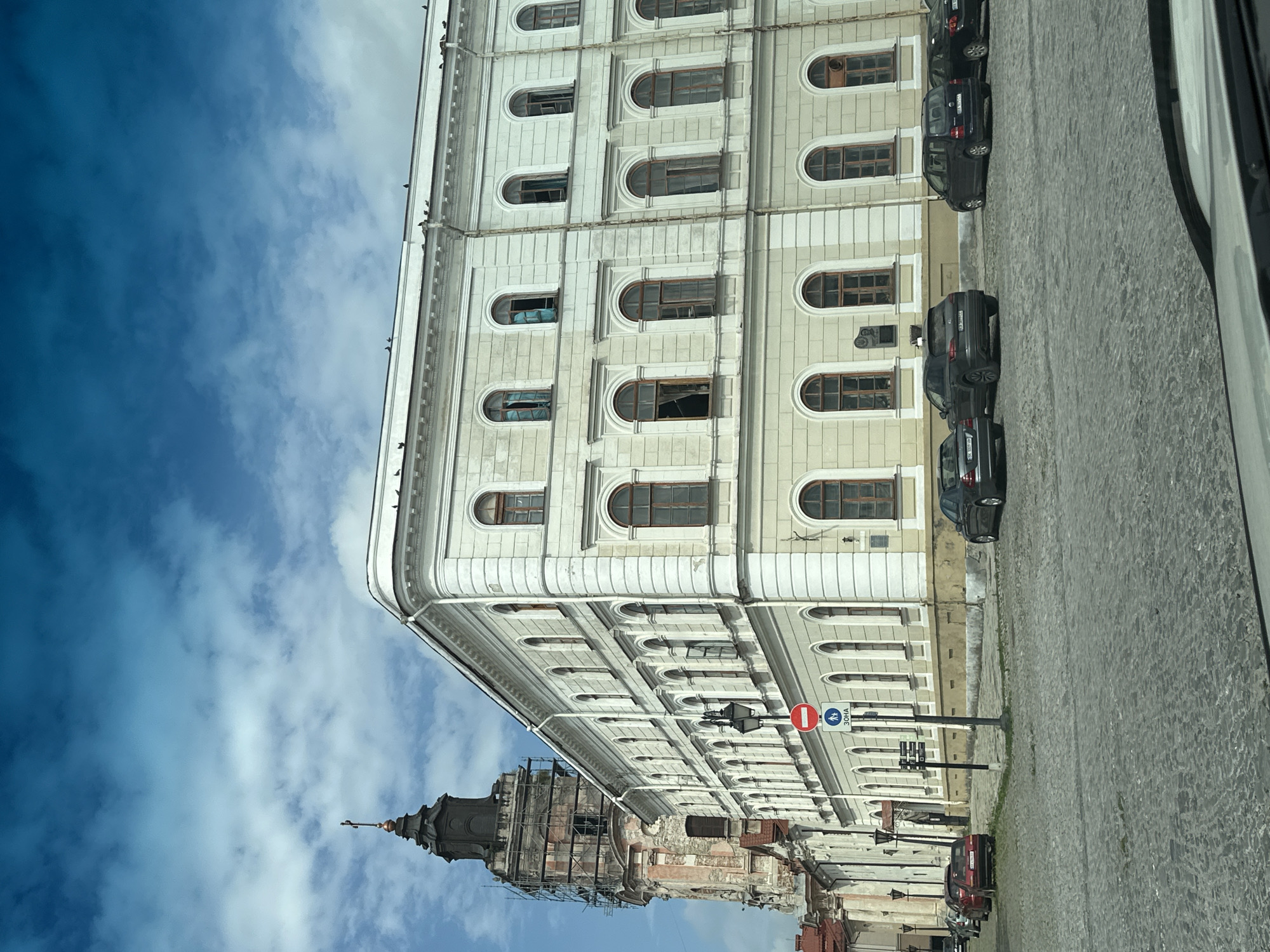 The image captures a view of the exterior of the Armenian Cathedral in Kamianets-Podilskyi, Ukraine. The cathedral's dominant architectural feature is its tall, dark tower, which is partially covered in scaffolding, indicating ongoing restoration work. The main building of the cathedral, visible to the right of the tower, is a light-colored, multi-story structure with numerous arched windows, many of which appear damaged or open. The facade of this building is adorned with classical architectural elements, including decorative moldings and pilasters.
In the foreground, a cobblestone street is visible, with several cars parked along the curb. These include a dark-colored SUV, a red hatchback, and a dark minivan. A street lamp and several traffic signs are also present on the street. The sky above is a vibrant blue, dotted with fluffy white clouds, suggesting fair weather. The overall scene depicts a section of an urban environment, with historical architecture and everyday elements like vehicles and street infrastructure.