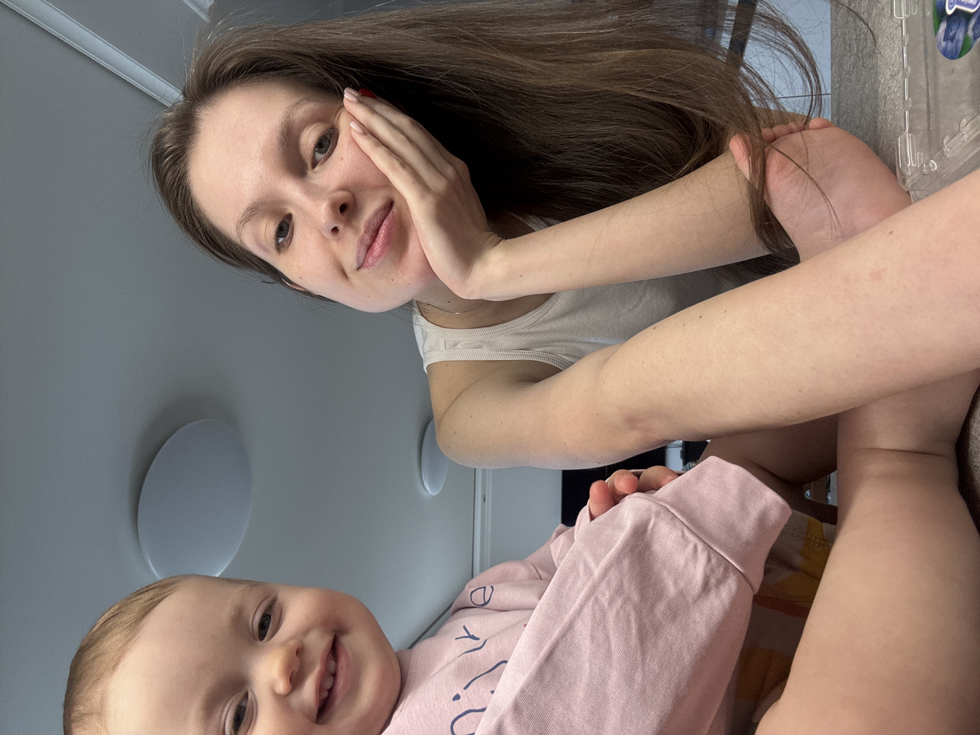 This image captures a joyful moment between a woman and a baby, likely mother and child, in an indoor setting in Moscow, Russia. The photo is taken from a low, rotated angle, suggesting a candid selfie.
The woman, positioned higher in the frame, has long, dark brown hair and fair skin. She is wearing a white tank top and has a gentle smile, with her chin resting on her right hand. Her fingernails are painted a subtle red. Her eyes are light in color, and she looks directly at the camera.
Below her, the baby has light brown hair and fair skin, sporting a wide, happy smile that reveals several small teeth. The baby is dressed in a light pink long-sleeved shirt, which has some indistinct dark blue text printed on it, possibly partially spelling out "LIVE" or a similar word.
The background shows a light-colored ceiling with a circular flush-mount light fixture. To the right of the woman, a clear plastic container is partially visible, with the word "BLUEBERRIES" clearly identifiable on its label, suggesting it holds fruit. Both subjects appear content and happy, creating an intimate and warm atmosphere. The lighting suggests it is daytime indoors.