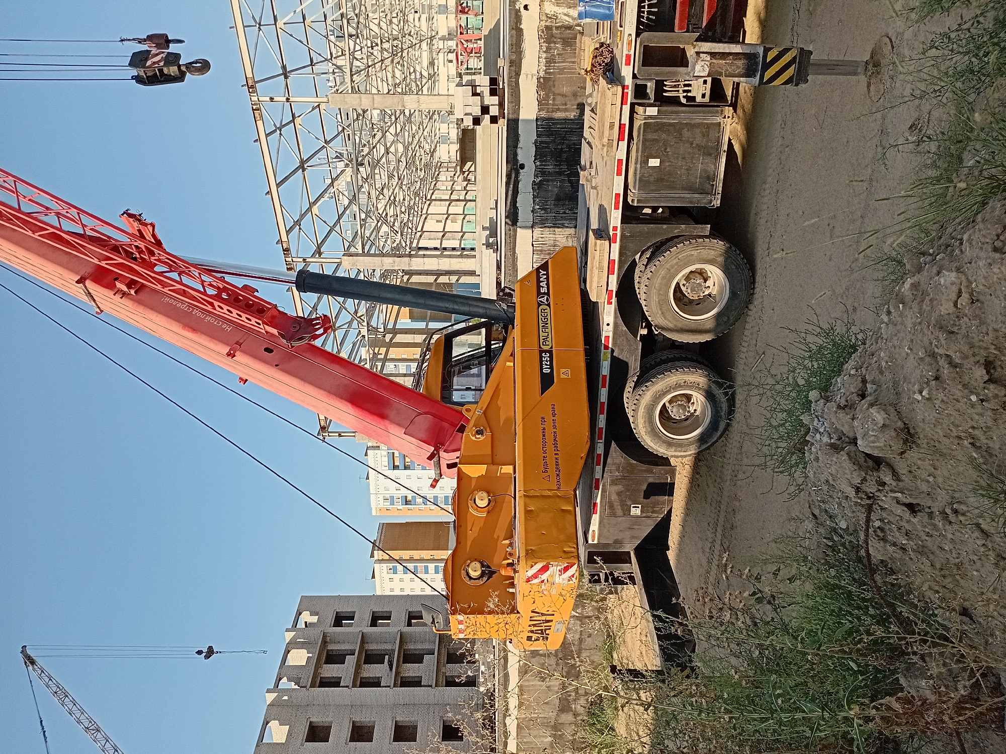 This is a medium shot, eye-level, outdoor photograph taken in the city of Semey, Kazakhstan. The image depicts a construction site with a large orange mobile crane as the main subject. The crane's boom extends upwards and to the right, with the hook mechanism visible. The crane is positioned on a dirt surface, with vegetation and piles of dirt in the foreground. In the background, a partially constructed building made of light-colored bricks is visible, along with a steel structure of a larger building under construction, and several multi-story apartment buildings further in the distance. The sky is clear and blue. The scene is brightly lit, suggesting it was taken during daylight hours. No people are visible in the image.