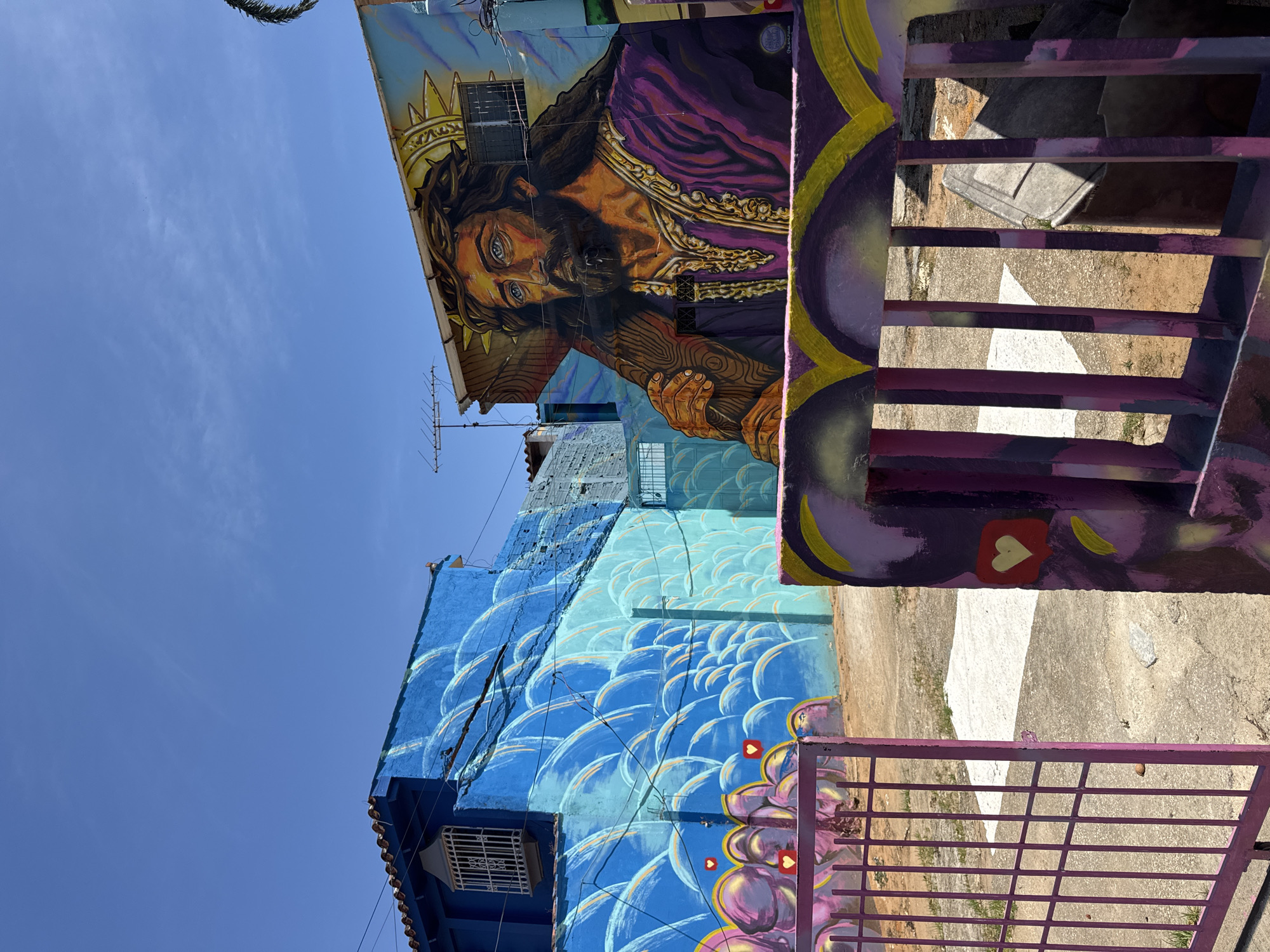 The image captures a vibrant street scene in La Asuncion, Venezuela, featuring multiple buildings extensively covered in colorful murals under a clear blue sky. The perspective looks upwards, showcasing the painted facades.
The most prominent subject is a large mural of Jesus Christ painted on the upper part of a building. He is depicted with dark, flowing hair and a beard, wearing a crown of thorns, and a rich purple robe with golden details. His blue eyes gaze intently, and he is shown holding a wooden cross. A golden halo or sunburst emanates from behind his head. On the roofline of this building, a television antenna is visible, and the building features windows with protective grates. A palm frond is just visible in the upper left corner against the sky.
Below this, on an adjacent or lower section of the same structure, another building is painted with an intricate blue mural. This design features swirling, wave-like, or scale-like patterns in varying shades of blue and light blue, creating a sense of movement.
In the foreground, on the right side, a sturdy metal gate or fence painted in a deep purple hue is visible. This gate is adorned with abstract purple and yellow designs, and notably, several small red heart icons with white outlines, resembling social media "like" buttons, are incorporated into the artwork. Through the bars of the gate, a dirt and concrete path is visible on the ground.
The scene is bathed in bright daylight, indicating a sunny and clear day. On the mural of Jesus, near his left shoulder (viewer's right), a faint artist's signature, "JUAN CARLOS," is visible, along with a circular logo.
