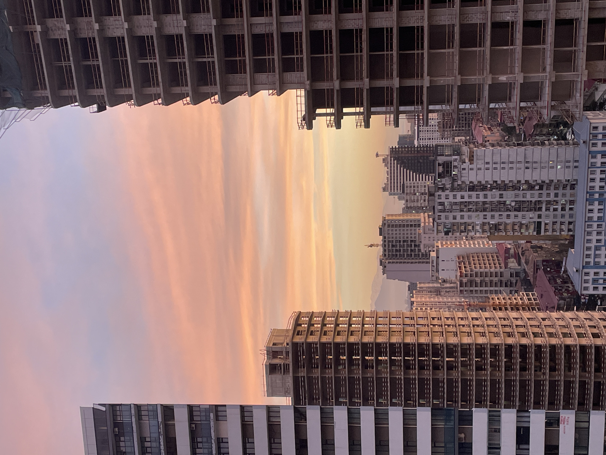 The image depicts an urban landscape at sunset in Batumi, Georgia. The foreground and top of the image are dominated by the incomplete skeletal structures of high-rise buildings under construction, revealing the grid-like pattern of their reinforced concrete frames. In the middle ground, existing buildings with white facades and grid-like windows line the horizon. The sunset casts a warm, pinkish-orange glow over the scene. A faint mountain range is visible in the distance. The image is upside down.