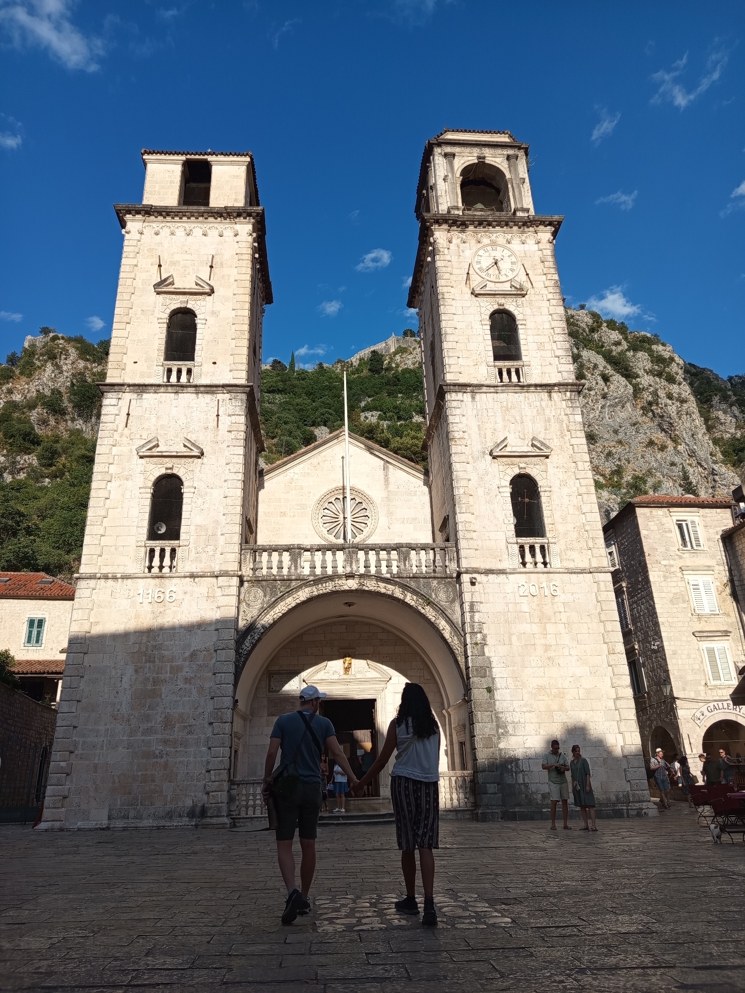 The image depicts the Square of the Cathedral in Kotor, Montenegro, with the iconic Cathedral of Saint Tryphon dominating the scene. The cathedral features two prominent bell towers, one of which displays a clock face. Carved into the stonework of the towers are the dates "1366" and "2016". The facade of the cathedral is adorned with an archway and a rose window. In the foreground, a couple is walking away from the camera, holding hands. Other individuals are present in the background, some interacting, while others appear to be tourists. The ground is paved with cobblestones, and the surrounding buildings are constructed of stone. The sky is a clear blue with scattered clouds, and a rocky, tree-covered mountain rises behind the cathedral. The scene is bathed in sunlight, casting distinct shadows.