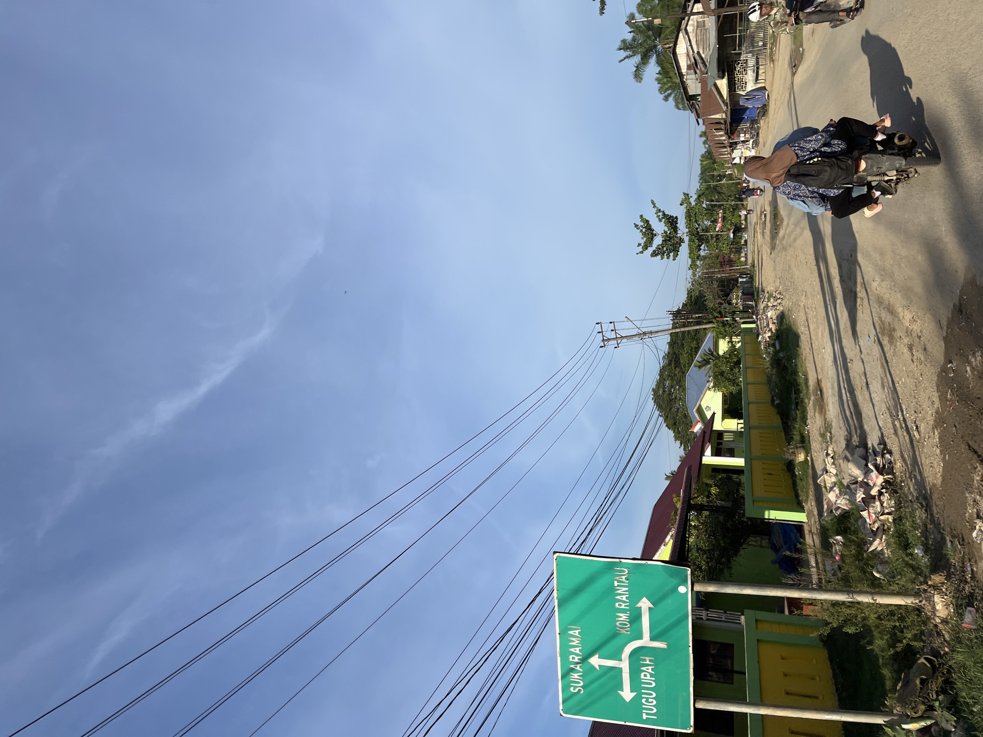 The image depicts a street scene in Alurcucur, Indonesia, under a bright, clear blue sky with scattered wispy clouds. A prominent green directional sign is visible on the left, indicating "SUKARAMAI" with an upward arrow, "TUGU UPAH" with a leftward arrow, and "KOM. RANTAU" with a rightward arrow. The sign is mounted on two metal poles. To the right of the sign, a dirt road stretches into the distance, lined with houses and shops on both sides. Several power lines are strung across the sky.
In the foreground, a dirt road is visible, with debris and small piles of rubble scattered along its edge, particularly near the sign. The road surface appears uneven and dusty.
Several people are present in the scene. Two individuals are riding a motorcycle in the middle of the road, facing away from the camera. The person in the back is wearing a hijab. Further down the road, two more motorcycles are visible with riders, and another person appears to be walking.
The buildings lining the street are varied in style and condition. Some appear to be houses with brightly colored walls (yellow and green are noticeable) and red or blue roofs. Others seem to be more modest shops or residences with corrugated metal roofs. Vegetation, including trees and palm trees, is present throughout the scene, adding to the tropical atmosphere. The lighting suggests it is daytime, likely late afternoon given the long shadows cast by the power lines and buildings.