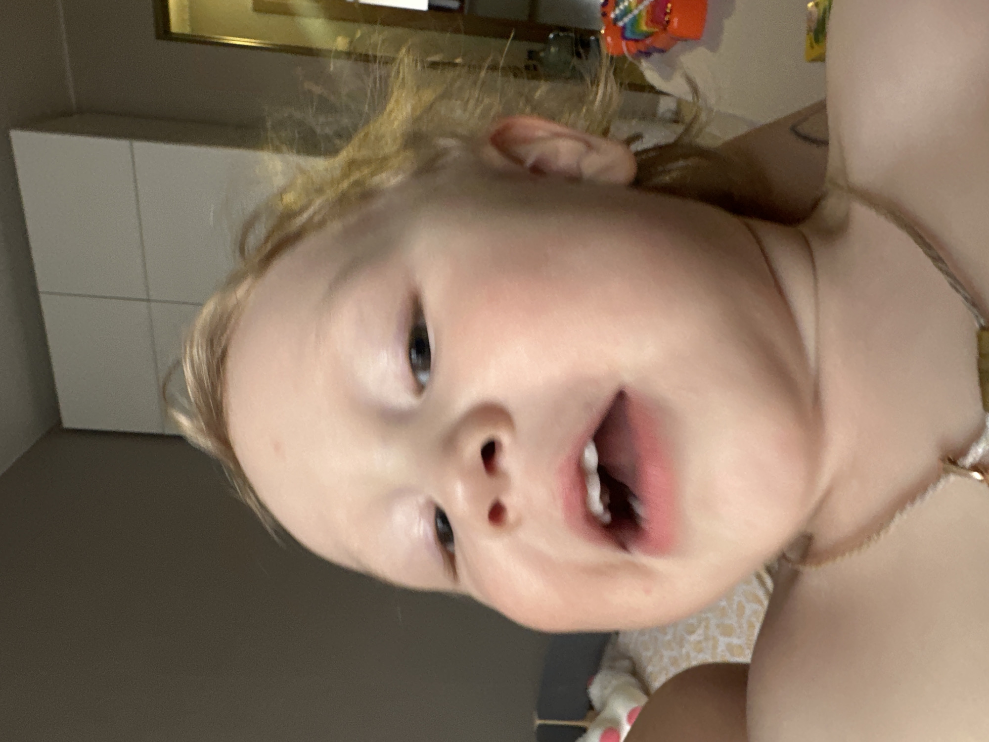 A close-up, rotated portrait shows a young child with fair skin and light, wispy hair, captured in an indoor setting. The image is oriented sideways, with the child's head tilted to the right. The child's face dominates the frame, looking directly at the viewer with wide, dark eyes. The mouth is open, revealing small teeth, suggesting an expression of surprise, playfulness, or mid-sound. A delicate, light-colored necklace is visible around the child's neck.
The background is out of focus, indicating a shallow depth of field. To the left of the child's head (which would be the top if the image were rotated upright), there is a white, paneled piece of furniture, likely a cabinet or wardrobe. Above this, a golden-framed object can be faintly seen. To the right, colorful, blurry shapes, possibly toys, and a patterned fabric, potentially bedding, are visible. The overall ambiance suggests a well-lit, cozy home environment, likely in Votkinsk, Russia. There is no visible text in the image.