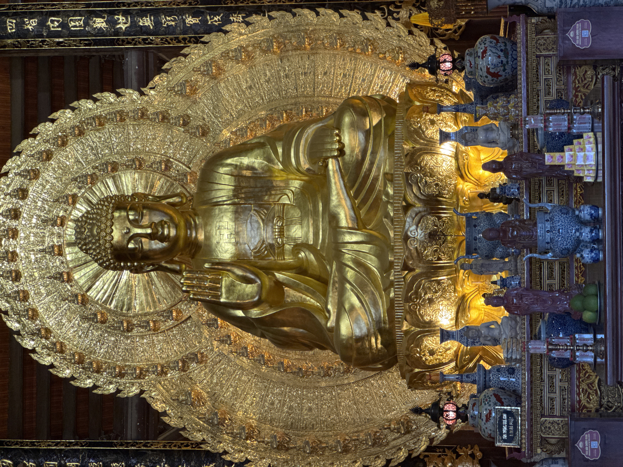 A full shot shows a large, golden statue of Buddha seated on a lotus throne. The statue is ornate, with intricate detailing on its robes and a radiating halo of smaller Buddha figures behind it. To the left and right of the main statue are tall, dark pillars with inscribed golden characters. In front of the Buddha statue, on a tiered altar, are various offerings and decorative items. These include several blue and white porcelain vases, small statues of other figures, a stack of what appear to be small boxes of incense or offerings, and some green fruit in a bowl. A red lantern hangs on the left side of the altar. The background consists of dark wooden beams and panels, typical of a temple or religious structure. The scene is dimly lit, with spotlights illuminating the golden Buddha statue and parts of the altar. This religious setting is located within the city of Tây Hoa Lư Ward, Vietnam.