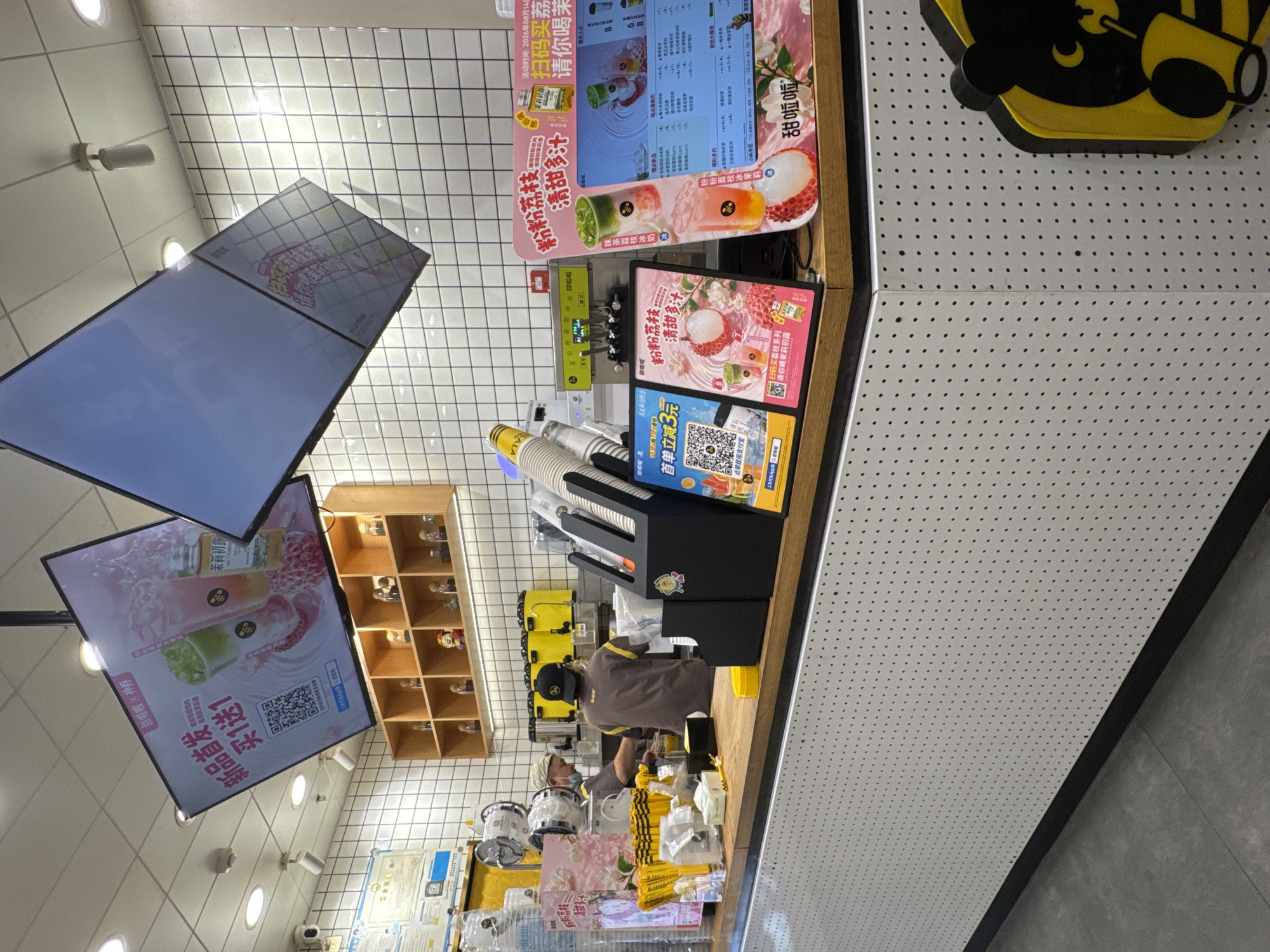 This image depicts the interior of a bubble tea shop, likely located in Shenyang City, China, as indicated by the presence of Mandarin Chinese text on various signage and displays. The shop features a bright, modern aesthetic with white tiled walls and a wooden counter. Two employees are visible behind the counter, one of whom is actively preparing drinks.
In the foreground, a wooden counter displays several promotional signs. One sign, prominently featuring lychee and green tea drinks, advertises "粉粉荔枝 清甜多汁" (Pink Lychee, Sweet and Juicy) and encourages customers to "扫码购买 请你喝" (Scan code to purchase, enjoy a drink). Another sign, partially visible on the left, shows a QR code and text related to a "首单立减3元" (First order minus 3 yuan) promotion. To the right, a large digital screen displays a pink-themed advertisement for new products, with text like "新品首发 买1送1" (New product launch, buy 1 get 1 free).
Behind the counter, a selection of beverage preparation equipment is visible, including yellow dispensers and stainless steel machinery. Stacks of paper cups and other drink-making supplies are neatly arranged. The overall environment suggests a busy and inviting establishment focused on promoting its beverages. The background shows shelves stocked with decorative items, and the ceiling has overhead lighting fixtures. The floor appears to be a neutral-toned tile.