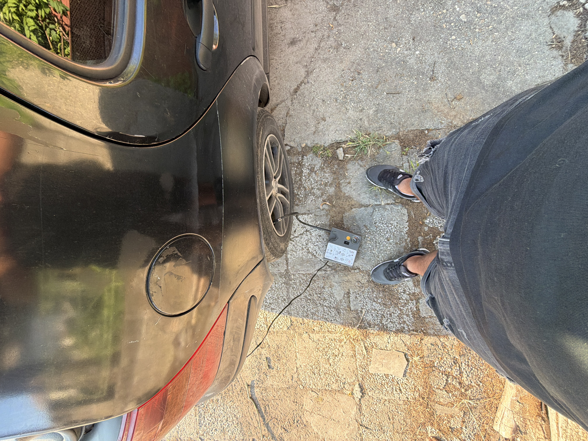The image captures a first-person, downward-looking perspective of an individual using an air compressor to inflate a car tire in Kalipetrovo, Bulgaria. The scene is set outdoors on a sunny day, with bright, direct sunlight creating distinct shadows.
The subject's lower body is visible, wearing dark jeans, possibly with some distressing or rips, and dark athletic shoes with white accents. They are standing on a ground surface that appears to be a mix of cracked asphalt, small stones, and patches of dry grass or weeds.
To the left, a black car, likely a hatchback, is partially visible. The rear passenger door, the rear wheel, and the fuel cap are prominent. The car's body shows some dirt and minor scratches. A portable air compressor, primarily grey with yellow/orange accents, is positioned on the ground next to the car's rear wheel. A black hose extends from the compressor, connecting directly to the valve of the car's tire, indicating the inflation process is underway.
The air compressor features a gauge and several symbolic icons on its surface, depicting various items that can be inflated, such as car tires, bicycle tires, and sports balls. While specific text is unreadable, these symbols likely correspond to operational instructions or pressure settings. The overall impression is one of practical car maintenance taking place in a rustic, outdoor environment.