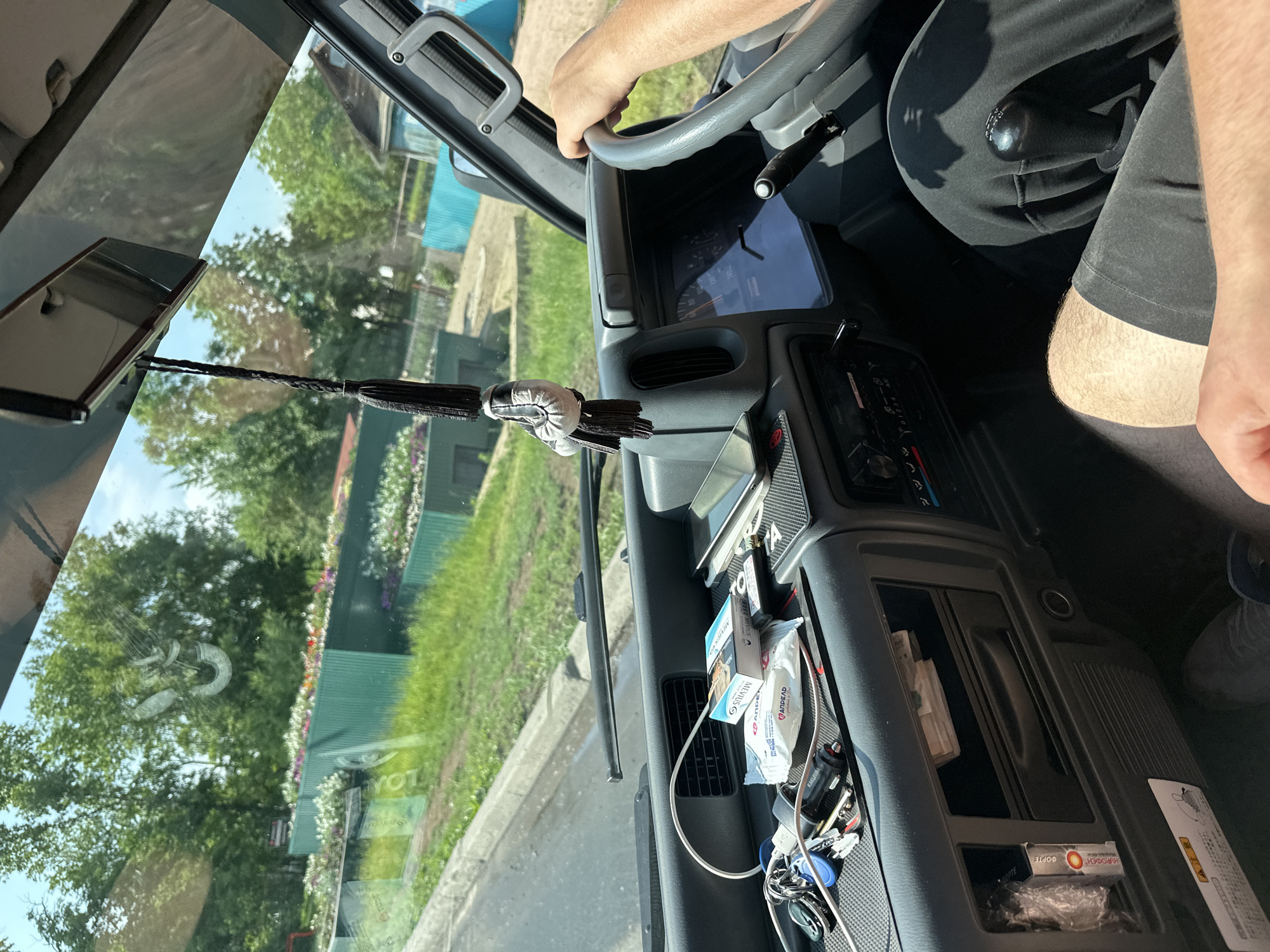 The image shows the interior of a vehicle, likely a van or truck, from the perspective of the driver or passenger. A hand is visible on the steering wheel, and the driver's leg is also in view. The dashboard is cluttered with various items, including a pack of cigarettes, a phone, keys, a car charger, and a package of wipes. A gear shift is positioned between the driver and passenger seats. Outside the vehicle, trees and some structures are visible through the windshield, suggesting an outdoor setting. The overall impression is of a personal vehicle in use. The image is taken from inside the cabin, looking forward. The lighting suggests it is daytime.