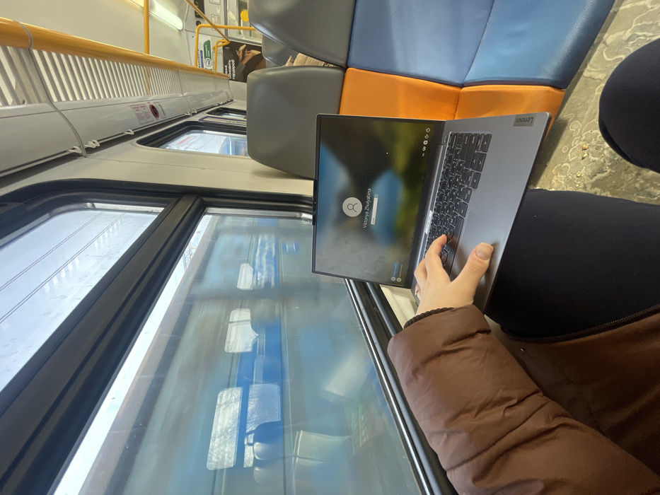 The image depicts the interior of a train carriage in the city of Бабеево, Russia. A person, whose left arm and hand are visible, is seated, wearing a brown puffer jacket and dark pants. A silver Lenovo laptop is open on their lap, with their left hand positioned over the keyboard, fingers resting on the keys. The laptop screen displays a login interface featuring a profile icon, the name "Viktoriya Zhulyabina," and a password input field, set against a blurred landscape background. Through the train's window on the left, a blurry blue train car is visible, indicating movement or proximity to another train. The interior of the carriage features rows of seats upholstered in grey, orange, and blue. Overhead luggage racks and fluorescent lighting are visible above. The floor has a light-colored, possibly patterned surface.