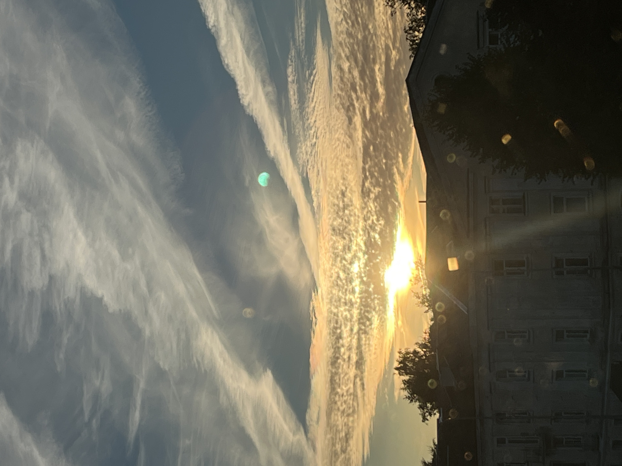 The image depicts a scene in Kohtla-Järve linn, Estonia, during what appears to be either sunrise or sunset, given the bright, low-lying sun illuminating the sky. The sky is a dynamic canvas of clouds, with wispy cirrus clouds streaking across a deeper blue. Lower down, the clouds are illuminated by the sun, creating a textured, golden-orange hue. There are two distinct luminous orbs in the sky; one is a pale yellow-white and slightly blurred, and the other is a vibrant cyan.
In the foreground, the upper portion of a multi-story building is visible, characterized by its light-colored facade and numerous rectangular windows, some of which are dark. A dark, dense canopy of trees partially obscures the building and the lower sky. Scattered throughout the image, particularly in the lower half and overlaid on the building and trees, are numerous out-of-focus circular light artifacts, likely lens flares or reflections from water droplets on the camera lens. The overall impression is an atmospheric, urban landscape under a dramatic sky.