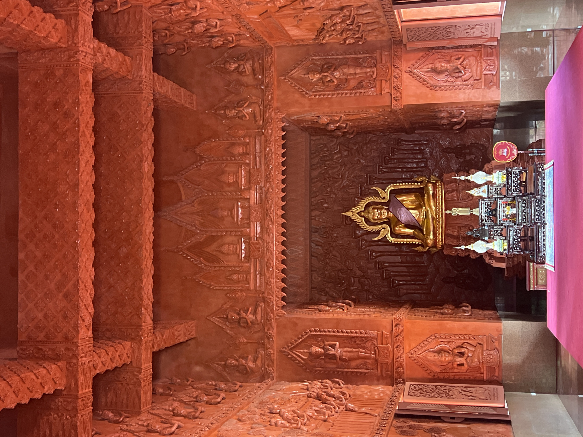 This is a frontal, eye-level, medium shot of the interior of a temple in Baan Hua Thanon, Thailand. The room is ornately decorated with intricate carvings in a reddish-orange hue, covering the walls, ceiling beams, and a recessed altar area. Dominating the altar is a large, golden Buddha statue seated in meditation, adorned with a gold crown and a purple sash draped over its shoulder. In front of the Buddha statue, a multi-tiered offering table holds various religious artifacts, including miniature statues and ornate containers. The floor in the foreground is covered with a bright pink carpet, and to the right of the altar, an open window reveals a glimpse of the exterior. The overall impression is one of opulent, spiritual grandeur.