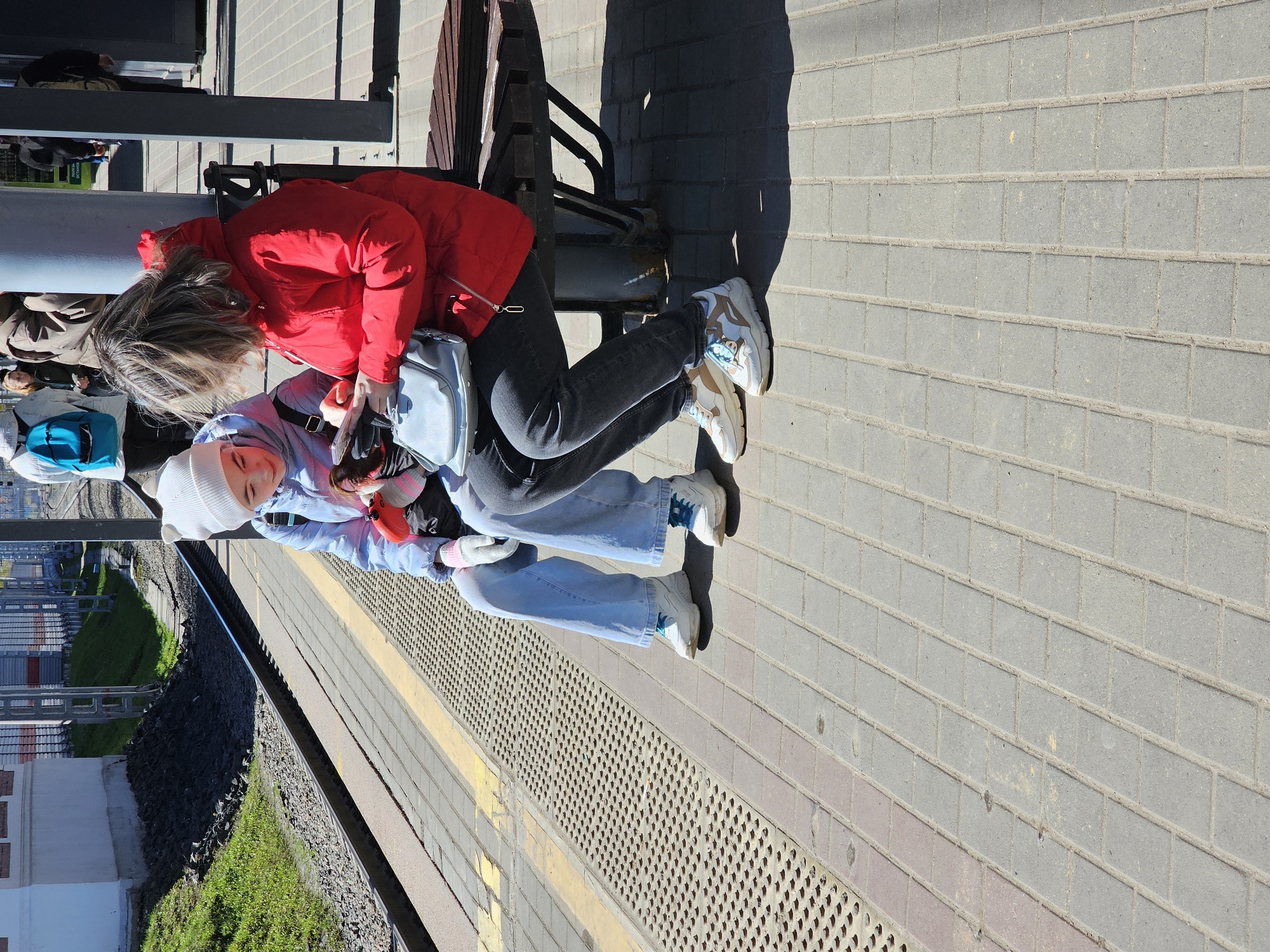 Two people are sitting on a bench at a train platform in Khimki, Russia. One person is wearing a red jacket and dark-colored jeans. The other person is wearing a light blue jacket, light blue jeans, and a white hat. There is a backpack near the bench. A train track with gravel and grass is visible next to the platform. The platform is paved with gray bricks.
