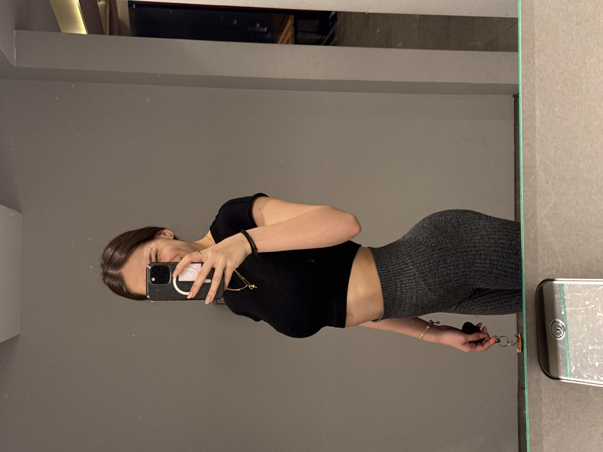 A woman stands in front of a mirror taking a selfie. She's wearing a black crop top and grey leggings. In her hand, she holds a set of keys. The background appears to be a bathroom with grey walls and fixtures. The location is likely a private residence or hotel room and no features indicating the city of Sofia are seen in the image.