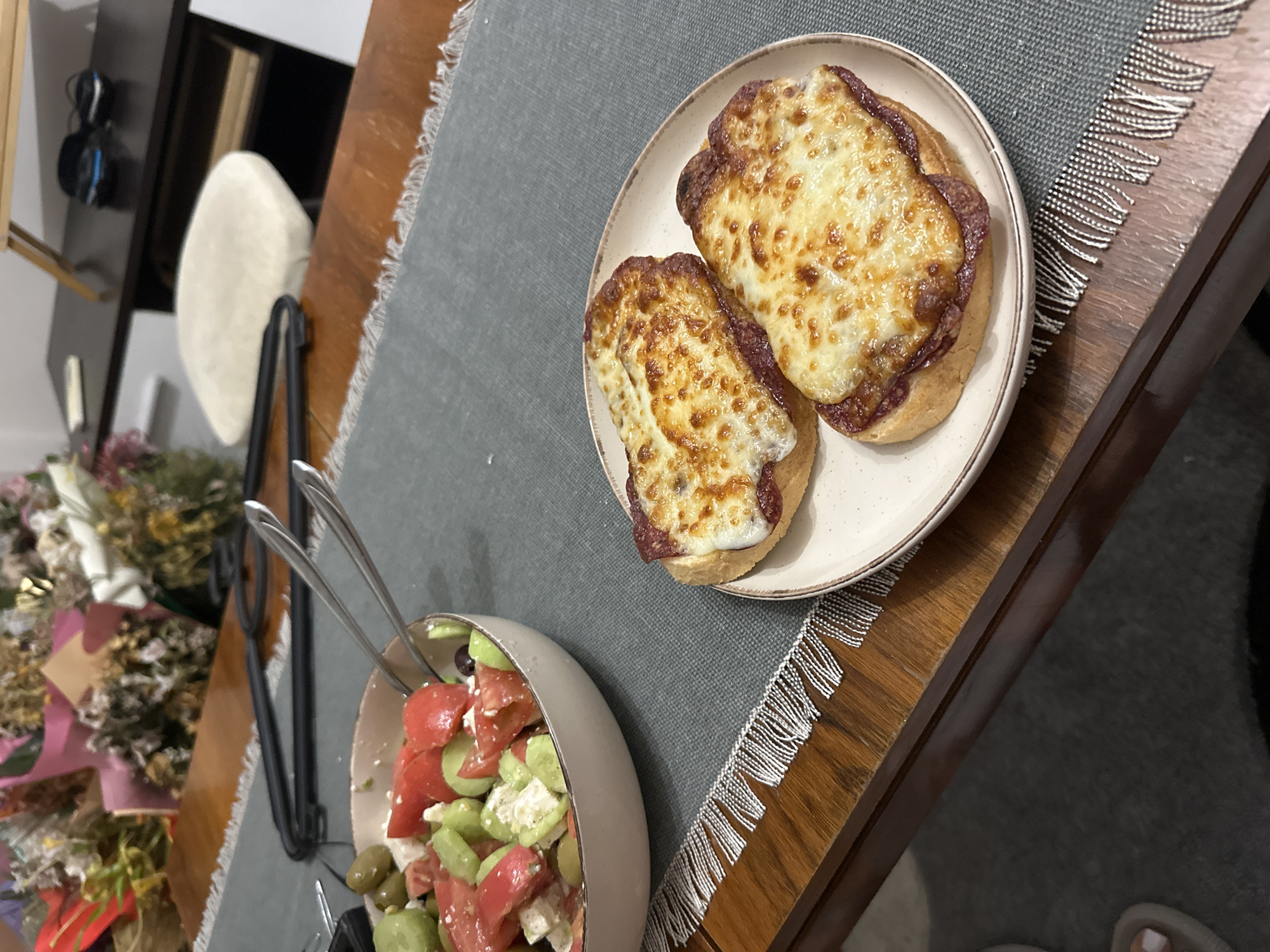This image captures an indoor dining scene in the city of Unknown, Unknown. The primary subjects are two food items placed on a dark gray woven table runner with white fringed edges, which rests on a polished wooden table. On the left, a light-colored bowl contains a salad composed of diced red tomatoes, sliced green cucumbers, cubed white feta cheese, and dark olives, with two metallic kitchen tongs partially submerged. To the right, on a round, speckled light-colored plate, are two pieces of open-faced toast. Each toast piece is topped with a reddish-brown spread and a layer of melted, golden-brown cheese, exhibiting distinct browning and bubbling, indicative of having been broiled or baked. In the blurred background to the left, multiple bouquets of dried flowers in various wrappers are visible. To the right, a dark brown low cabinet or console table is present, with a light-colored cushion or stool and a pair of dark objects (possibly slippers or shoes) on top of it. A wooden stand is partially visible at the far top right. The bottom left corner shows a partial view of a foot with white painted toenails, resting on a dark carpeted floor, suggesting a person is seated at the table.