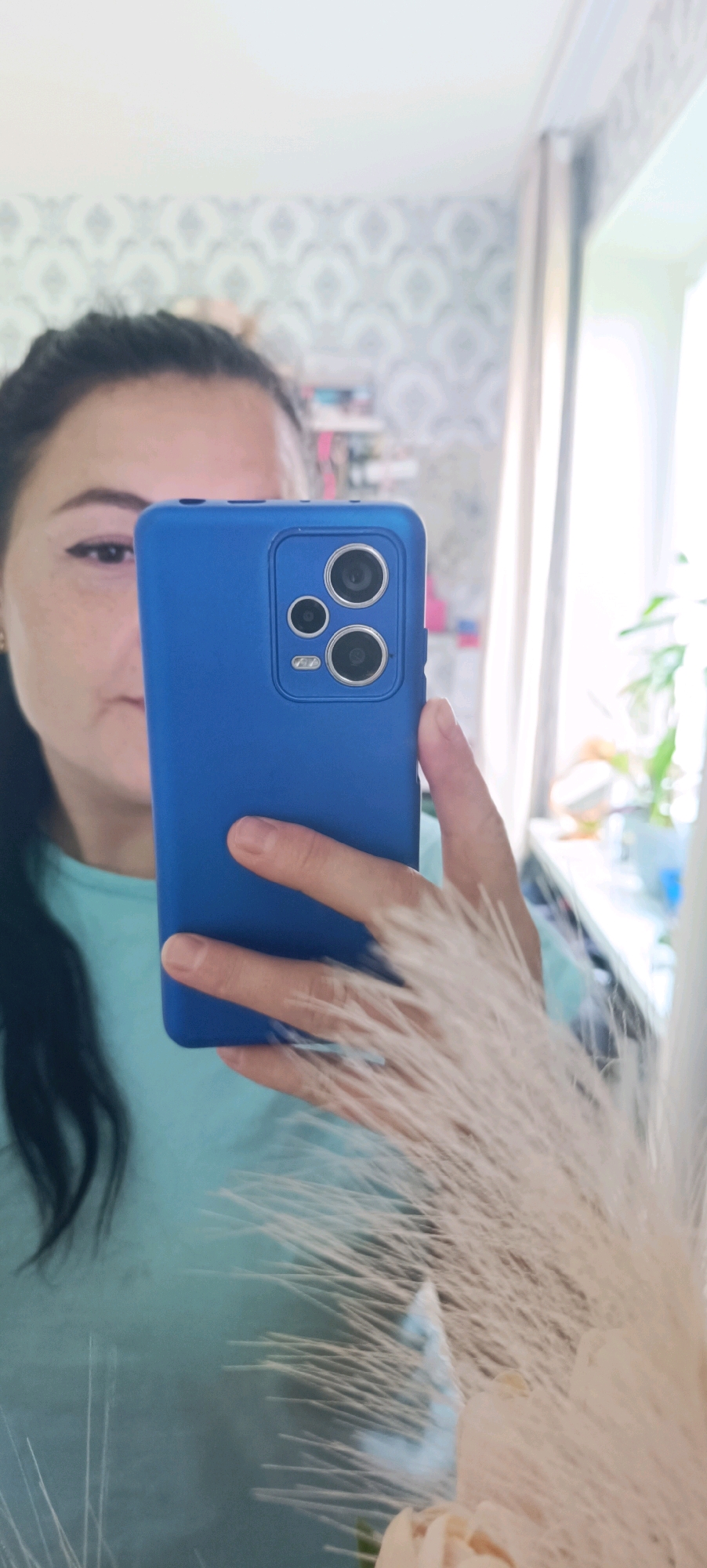 A woman is taking a selfie in a mirror, holding a blue smartphone. She has dark hair pulled back and is wearing a light blue t-shirt. The background appears to be a room with patterned wallpaper and a window with sunlight streaming in. There are plants visible through the window. The image is focused on the woman's face and the phone.