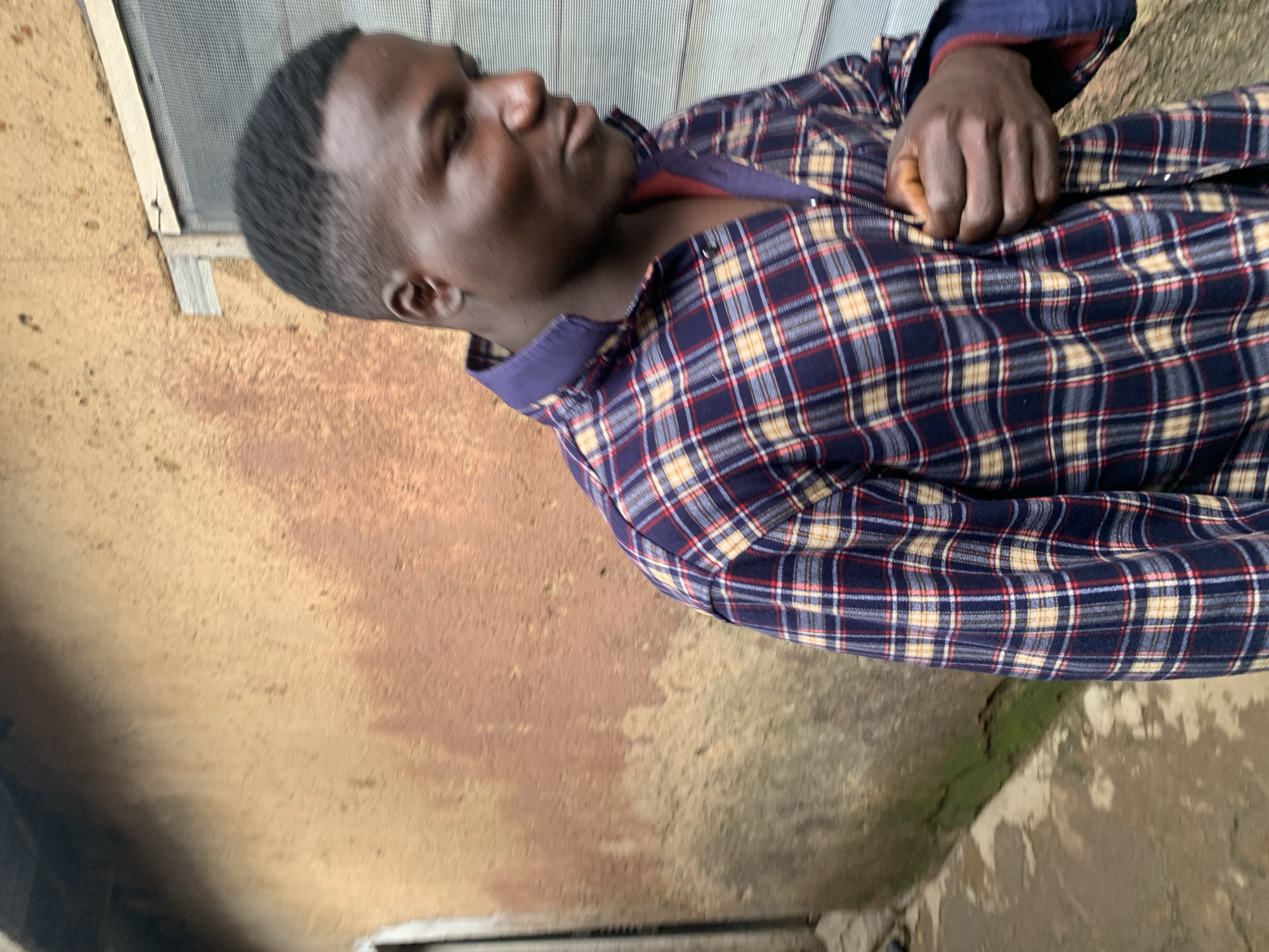 A man of African descent, with a short, faded haircut, stands in an outdoor urban setting in Jos, Nigeria. He wears a long-sleeved plaid shirt predominantly dark blue, with intersecting lines of beige/yellow and red forming the pattern. The shirt is partially unbuttoned at the top, revealing a dark-colored undershirt or inner lining, and its collar is a solid dark blue. His right hand is clasped around the front of his shirt, near the chest area. His head is angled downwards and to his right, with his gaze directed away from the camera. The background features a textured, weathered wall exhibiting shades of brown, reddish-brown, and beige, with areas of discoloration. To the right, a screened window with a light-colored wooden frame is visible. The ground is a light-toned paved surface, showing some dark and green discoloration along the base of the wall.