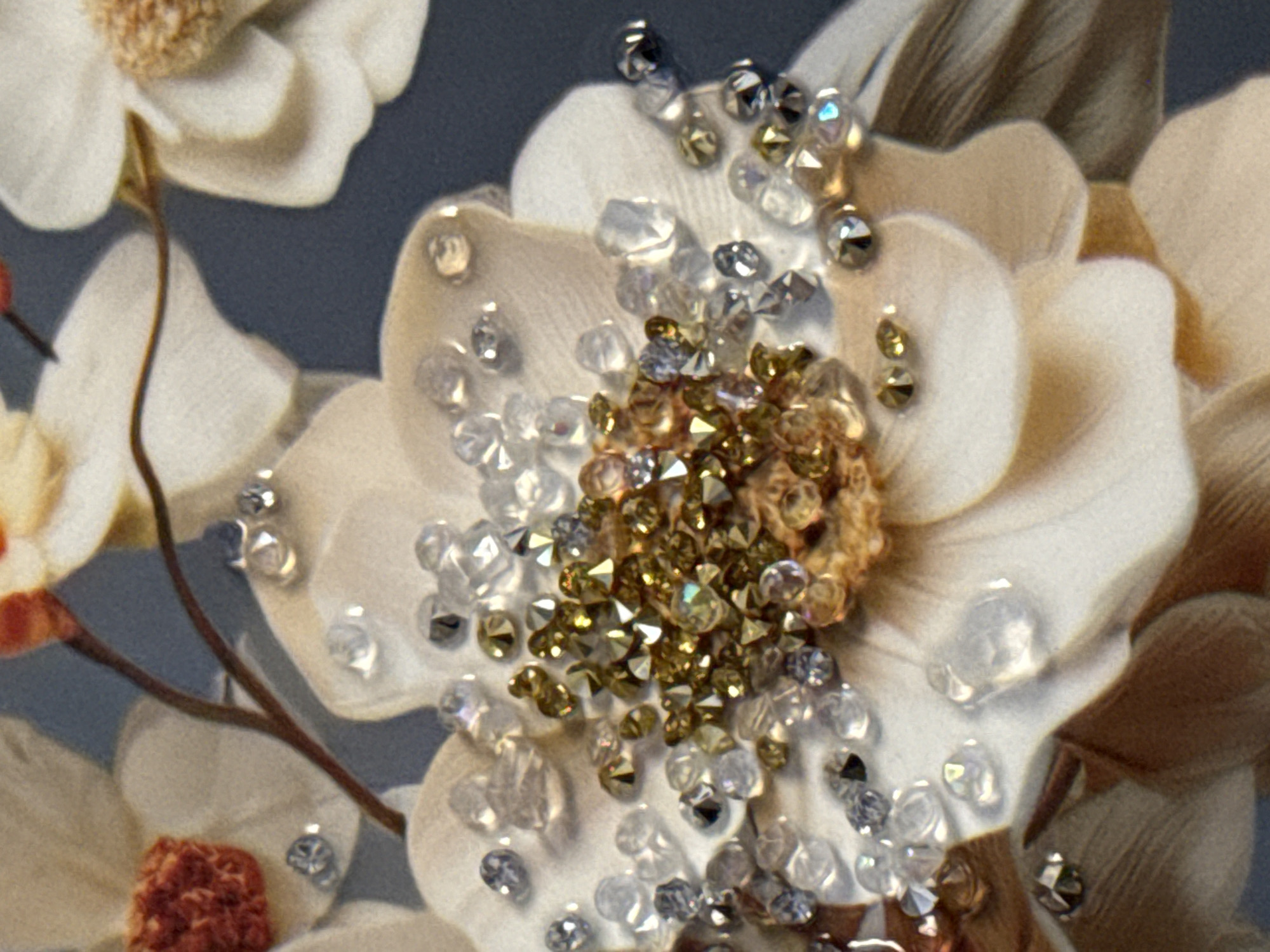 The image is a close-up, high-angle shot of a decorative floral arrangement, likely an artificial flower or a piece of artwork, embellished with numerous clear and gold faceted beads. The petals of the flower are rendered in shades of off-white and pale beige, with subtle shading that gives them a soft, layered appearance. Some of the petals have a textured, almost fabric-like quality.
The central part of the flower is densely clustered with the beads, creating a sparkling, jewel-like effect. The beads vary in size and shape, with some being round and others diamond-shaped. The clear beads reflect light, while the gold beads have a warm, metallic sheen. Scattered amongst the clear beads are some darker, possibly grey or deep purple, beads, adding contrast.
There are hints of other elements in the background, including what appears to be a brown stem or branch and possibly other flowers. The background itself is a dark, muted blue-grey color, which makes the details of the flower and its embellishments stand out prominently.
The image does not contain any identifiable landmarks, people, or actions, and therefore cannot be used to determine a specific location within the city of Tbilisi, Georgia. It is a detail-oriented photograph focused solely on the texture, color, and embellishments of the floral subject.