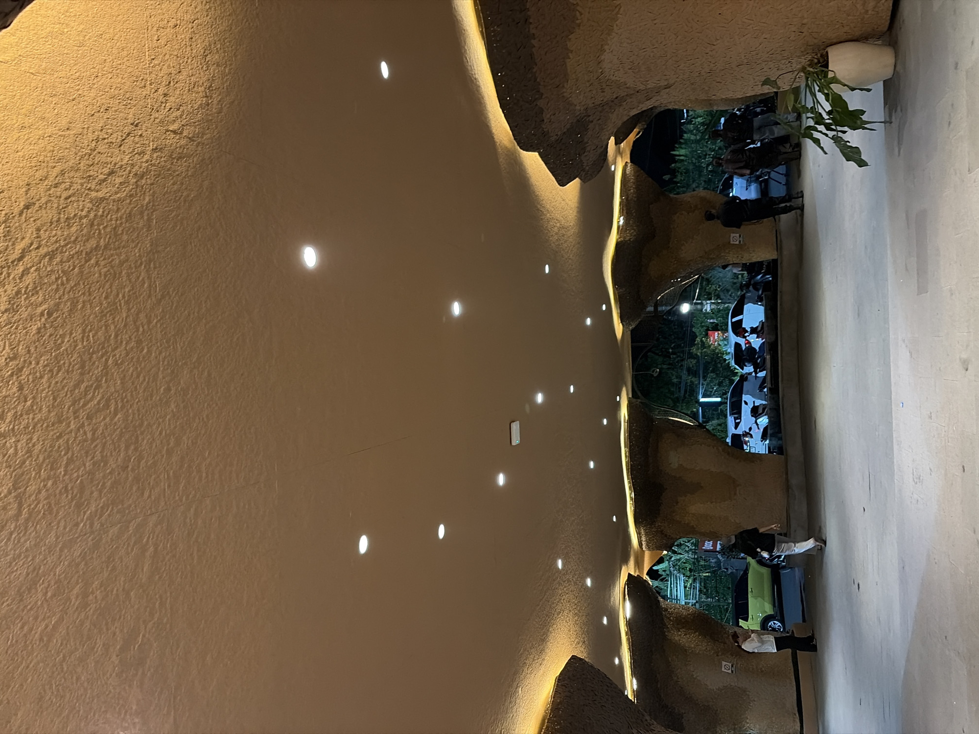 The image shows an outdoor area with a distinctive cave-like architectural overhang, illuminated by warm yellow lights along its edges. The ceiling of the overhang is a textured, light brown surface, with numerous circular lights embedded within it. The overhang structure itself has rough, textured surfaces resembling rock formations. In the background, through openings in the overhang, cars and people are visible. One car, a light green SUV, is parked on the left, and several other white cars and motorcycles are visible further back. Several individuals are walking or standing in the paved area beneath the overhang. On the right side of the image, a large potted plant with green leaves sits on the ground. The scene appears to be at night or dusk, given the artificial lighting and the darkness in the background. The overall impression is of an entrance or walkway to a building or attraction designed with a naturalistic, organic aesthetic. This location is identified as being in the Ubud District, Indonesia.