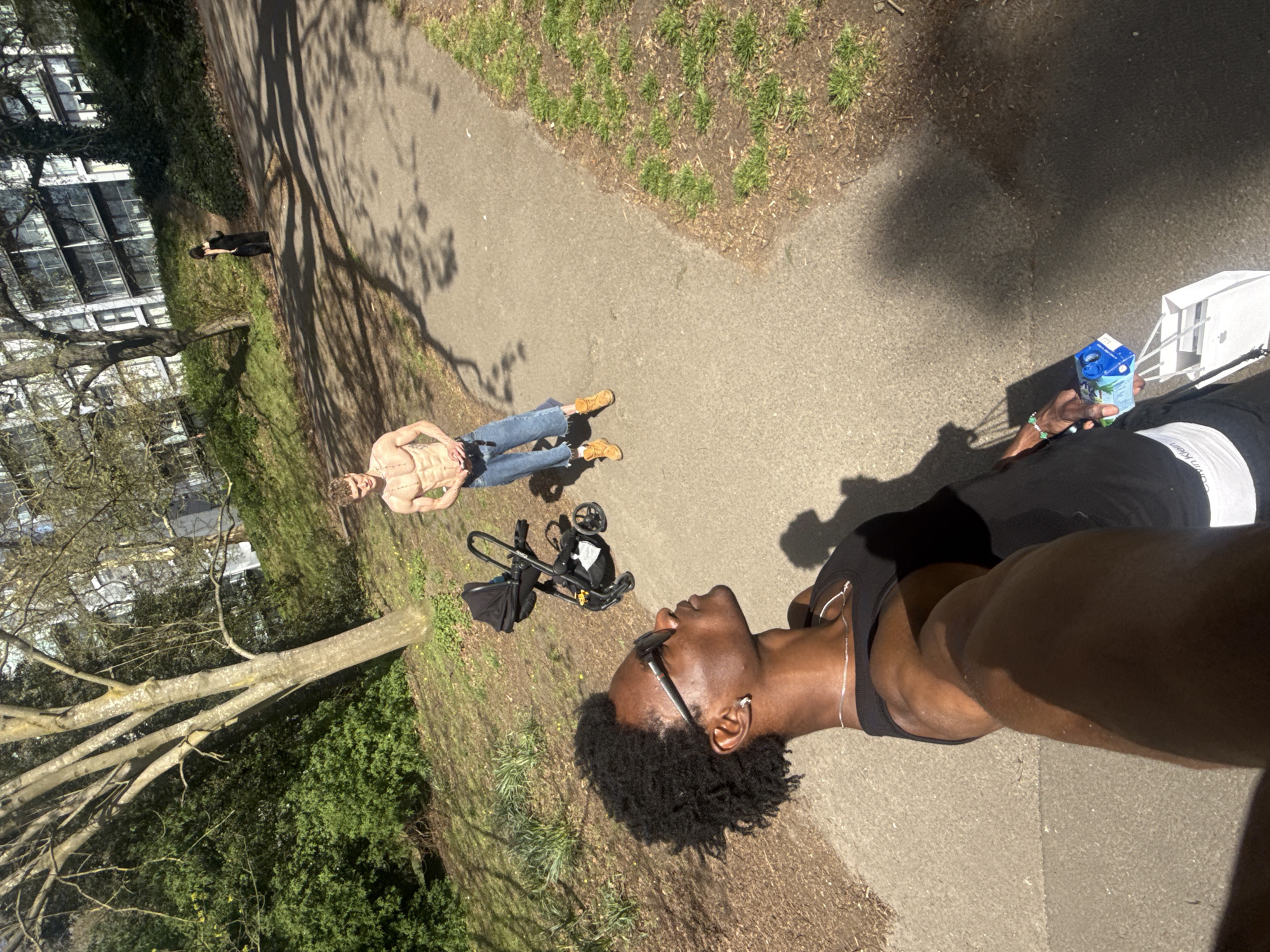 The image shows a scene in a park, possibly in London, United Kingdom. Two individuals are visible. One is lying on the ground in the distance, wearing jeans and tan shoes. Closer to the camera, another individual is also lying on the ground, holding a carton. A stroller and black bag are near the first person. There are trees and grass surrounding the paved pathway. Buildings are visible in the background. The camera appears to be close to the second individual's face.