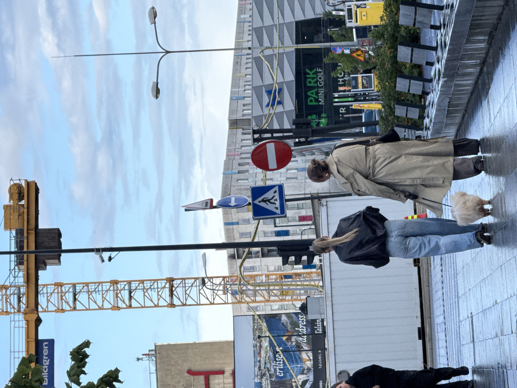 In Tallinn, Estonia, two women and a small, fluffy dog are walking on a wet, tiled sidewalk under a cloudy sky. The woman on the left wears a black jacket and blue jeans, and her hair is pulled back into a ponytail. The woman on the right is dressed in a tan trench coat and dark pants. The dog, which appears to be a bichon frise or similar breed, is light-colored and walking beside the woman in the trench coat.
The background features a construction crane, a tall building with a red "H" visible, a billboard advertising "erilise d'address Talsinki.e" and "Talsin," and modern buildings with geometric patterns on their facades. There are street signs, including a pedestrian crossing sign, a "no entry" sign, and directional arrows. A street light with two lamps is also visible. Further in the background, there's a sign that reads "PARK MINI GOLF" and a yellow gas station dispenser. The overall scene suggests an urban environment, possibly in a commercial or residential area undergoing development.