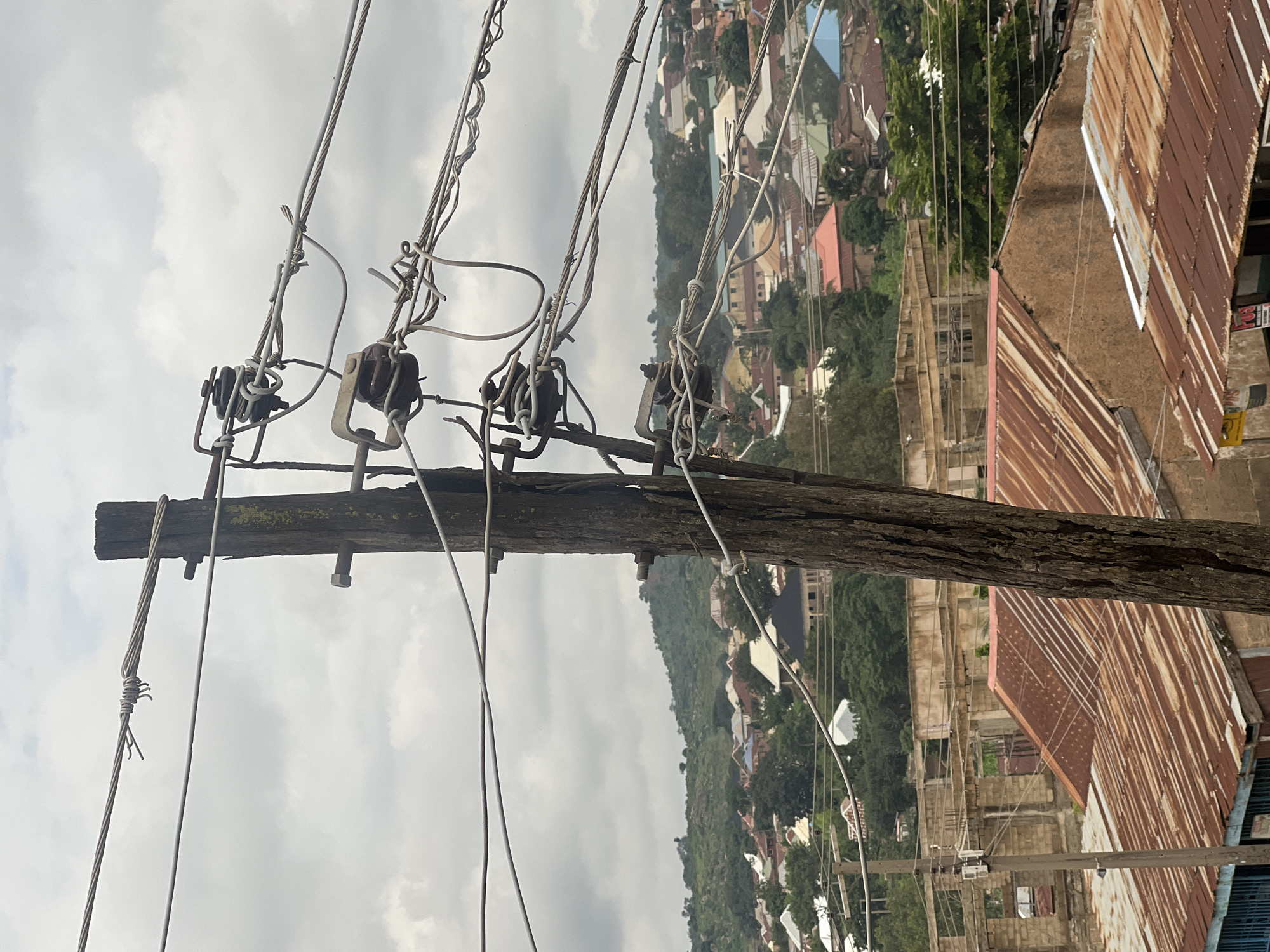 The image depicts a close-up view of a wooden utility pole with multiple wires attached, set against a backdrop of a city in Jos, Nigeria. The pole, weathered and vertical, is positioned in the foreground and is partially obscured by the wires. These wires, varying in thickness and condition, are strung across the frame and are attached to the pole via ceramic insulators. Some of the wires appear twisted or kinked, suggesting they may be old or damaged.
In the mid-ground and background, the urban landscape of Jos unfolds. Numerous buildings with corrugated iron roofs, in shades of red, brown, and grey, are densely packed together. Some structures appear partially constructed or in disrepair, with exposed brickwork and concrete frameworks. The city sprawls across rolling hills, dotted with lush green trees. The sky above is overcast with grey clouds, indicating a possibly humid or rainy day. There are no visible people or animals in the frame. The overall scene conveys a sense of a developing or established urban environment with visible signs of infrastructure.