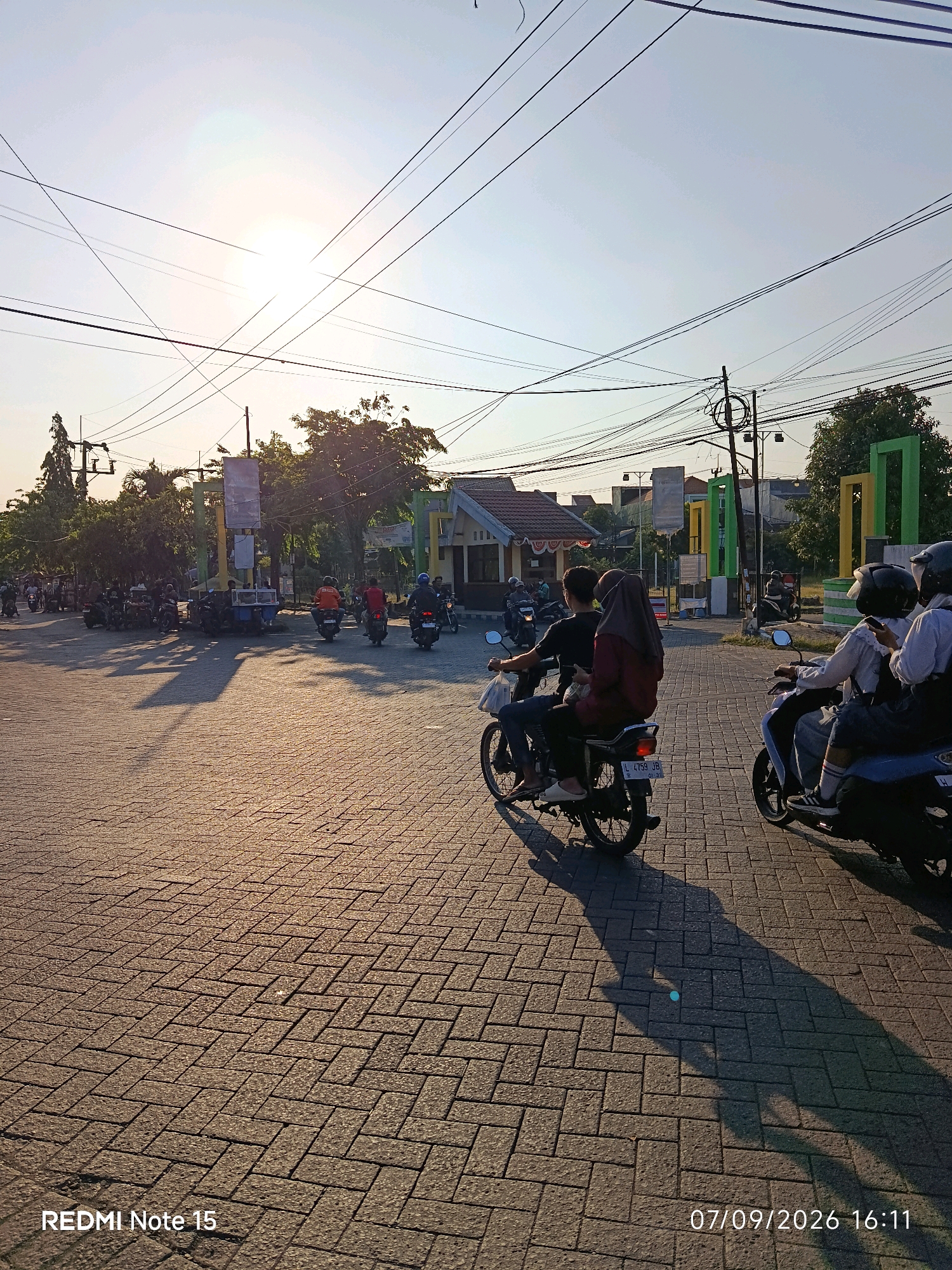In the city of Keboansikep, Indonesia, the image captures a street scene during what appears to be the late afternoon or early evening, as indicated by the long shadows cast by the sun. The ground is paved with interlocking brick pavers arranged in a herringbone pattern, reflecting the warm, low-angled sunlight. Overhead, a dense network of power lines crisscrosses the sky, silhouetted against a pale sky with the sun partially obscured.
Several individuals are seen riding motorcycles. In the foreground, two motorcycles are prominently featured. One carries two people, a male driver and a female passenger wearing a hijab and a reddish-brown top. The second motorcycle in the immediate foreground is partially obscured, with at least two people visible, one wearing a white long-sleeved shirt and the other also in white. Further in the background, more motorcycles and riders are present, suggesting a busy street.
On the right side of the image, a structure with a beige roof and supporting columns, some painted green and yellow, is visible, possibly marking an entrance or a building facade. Trees with dense foliage are scattered throughout the background, adding to the urban greenery. The overall impression is a snapshot of daily life in a Southeast Asian urban environment, with the presence of many motorcycles indicating a common mode of transportation. The timestamp in the lower right corner reads "07/09/2026 16:11". The "REDMI Note 15" watermark is present in the lower left corner.