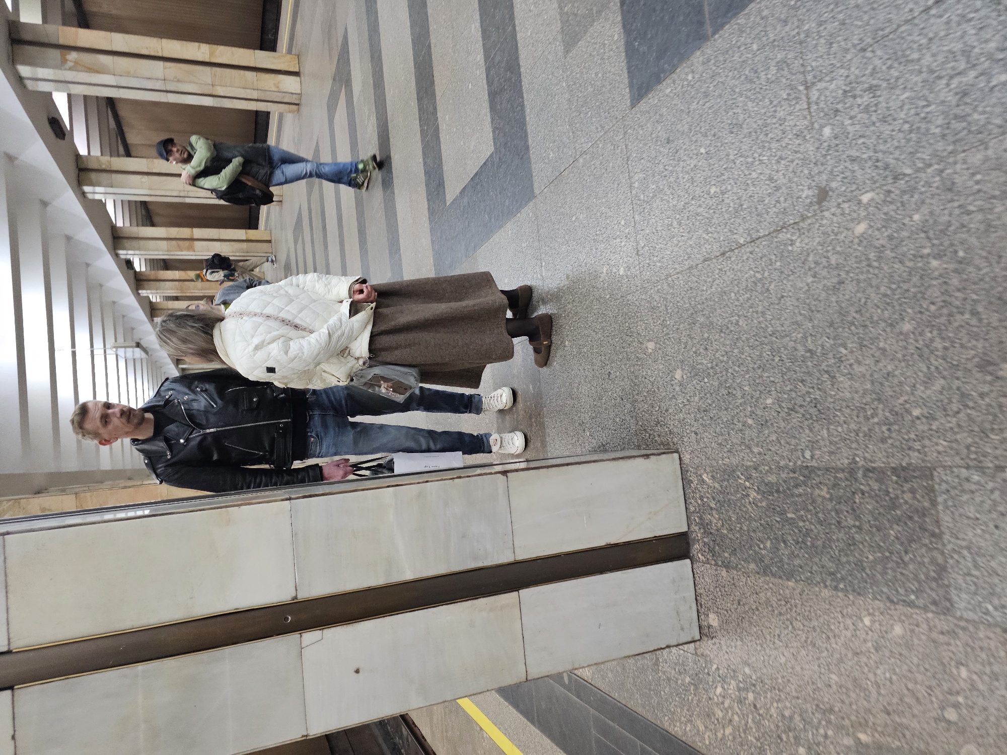 The image depicts a subway station, identifiable by the tiled walls, escalator, and striped floor pattern. Three people are visible. A man in a black leather jacket and jeans stands near the escalator; an older woman in a white quilted jacket and a brown skirt stands next to him. In the background, a younger man in a green jacket and jeans walks in the opposite direction. The location is likely in Khimki, Russia, based on contextual knowledge.