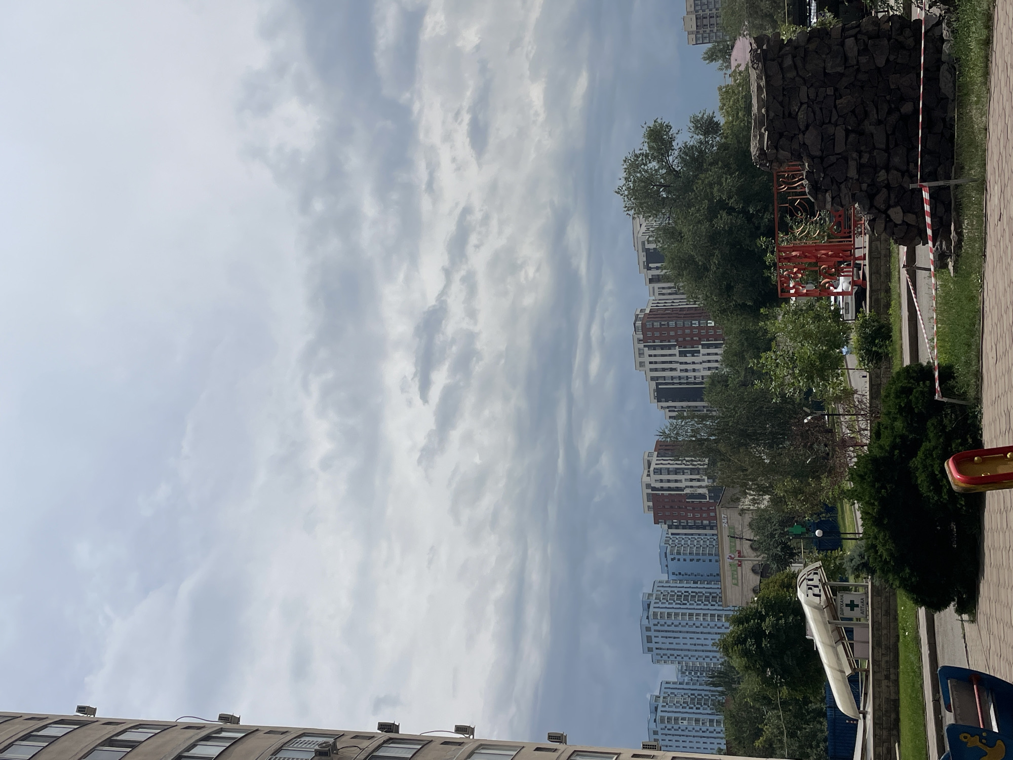 The image captures an overcast sky above a residential area in Almaty, Kazakhstan. In the foreground, a playground structure is partially visible on the left, and a low stone monument, cordoned off with red and white tape, stands on the right. Behind these elements, a street runs horizontally with a bus stop shelter marked "АПТЕКА" and a cross symbol on its side. Further back, a dense line of trees partially obscures a row of modern, multi-story buildings with light-colored facades and numerous windows. The sky is filled with gray, textured clouds.