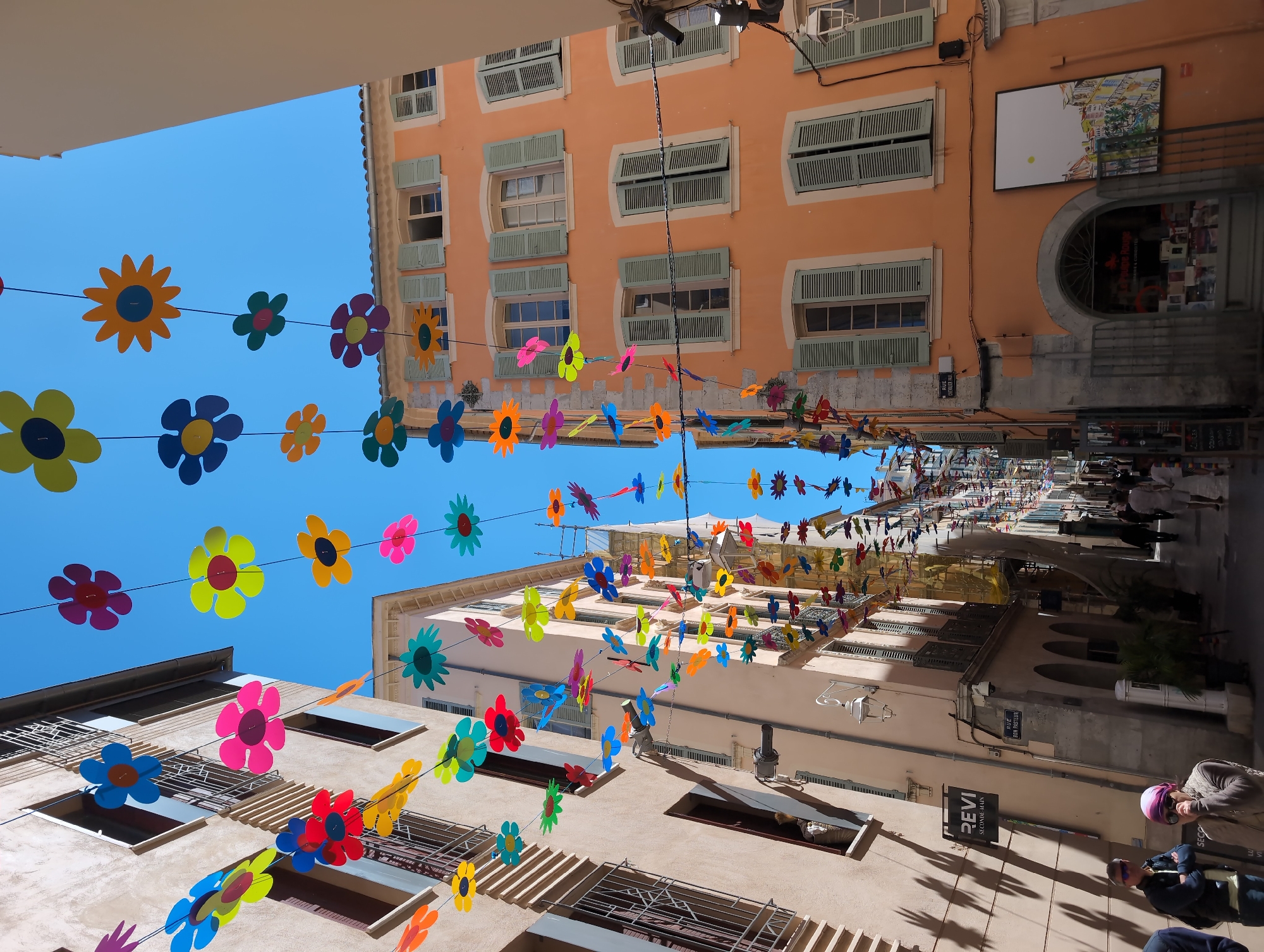 A street scene in Toulon, France, is captured from a low-angle perspective, looking up at buildings adorned with colorful flower decorations strung across the street. The architecture is characterized by buildings in shades of peach and cream, with windows featuring green shutters. The sky is a clear blue. People can be seen walking along the street, and signs indicate the location is "Rue Bon Pasteur" with a business named "PEVI" in view. Sunlight casts shadows on the buildings and street.