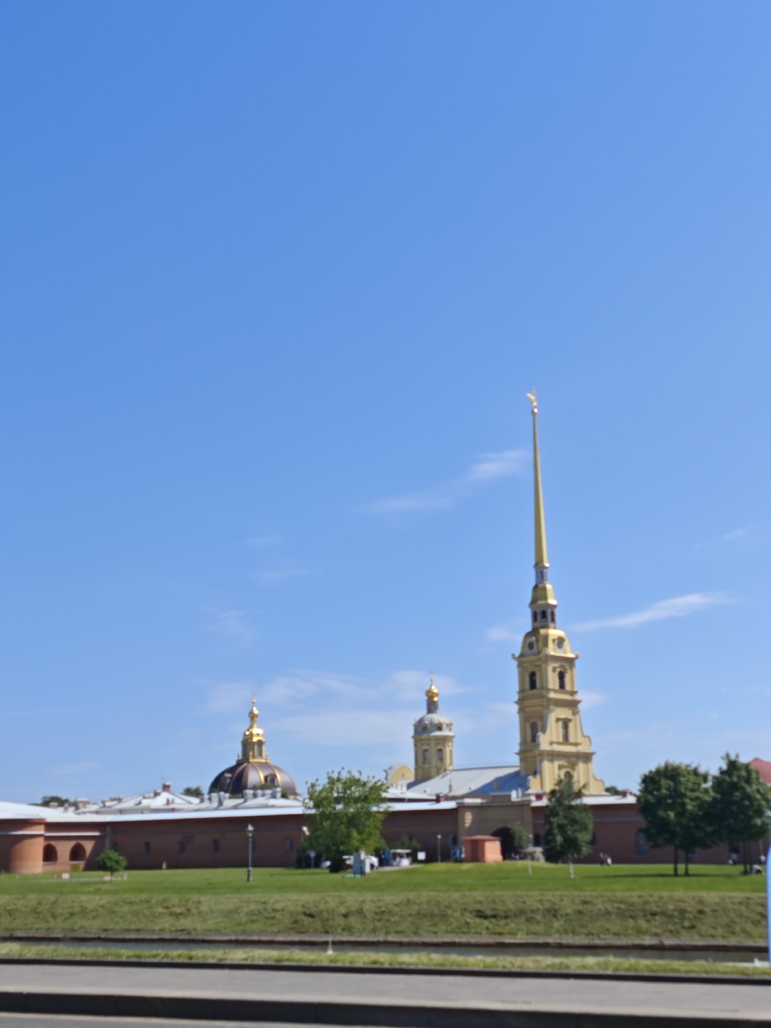 The image shows a wide shot of the Peter and Paul Fortress in Saint Petersburg, Russia, on a clear, sunny day. The fortress, made of red brick, features several prominent structures with golden domes and spires. The tallest structure is a yellow bell tower with a golden spire topped by an angel. To its left is a large building with a dark dome also topped with a golden spire. In the foreground, a green lawn stretches towards a body of water, and a paved walkway runs along the water's edge. Several trees are visible on the lawn. The sky is a vibrant, cloudless blue. A few distant figures can be seen on the grounds of the fortress, but they are not distinct.