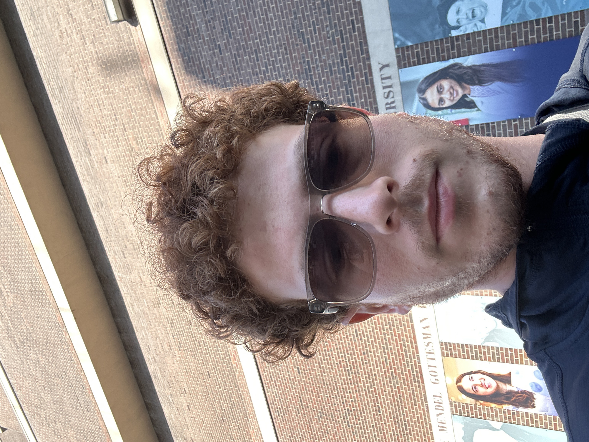 A man with curly brown hair and a beard stands in front of a brick building with banners of people. The banners include the words "University" and "Mendel Gottesman". This indicates the location is likely Yeshiva University in New York City. The man is wearing sunglasses and a dark blue shirt. He appears to be taking a selfie. The image has been rotated.