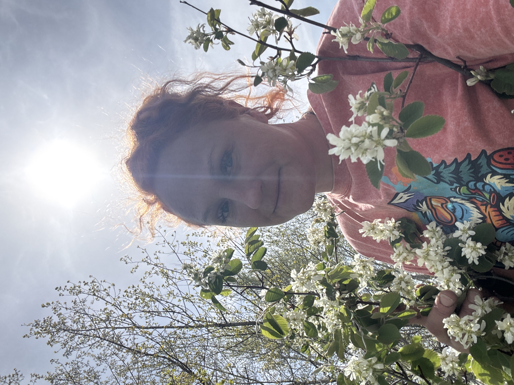 A low-angle medium shot shows a fair-skinned woman with reddish-auburn hair, wearing a pink t-shirt with a graphic design on the shoulder and torso, looking down at the camera. The woman's face is somewhat obscured by strong sunlight coming from the left of the frame. She appears to be holding branches of a flowering tree or shrub with small white flowers. The background shows branches of a tree or shrub, and glimpses of a bright sky. Location cannot be verified, and is presumed to be somewhere other than Chaikovsky, Russia.