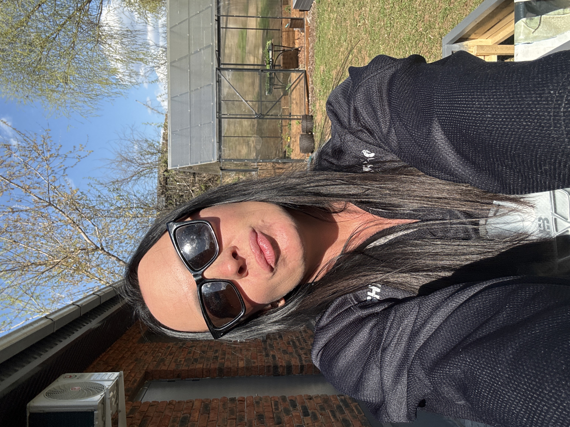 A woman wearing sunglasses and a black shirt is pictured outdoors. In the background, there is a greenhouse, a brick wall, a tree, and a blue sky. The location is potentially in Chaikovsky, Russia.