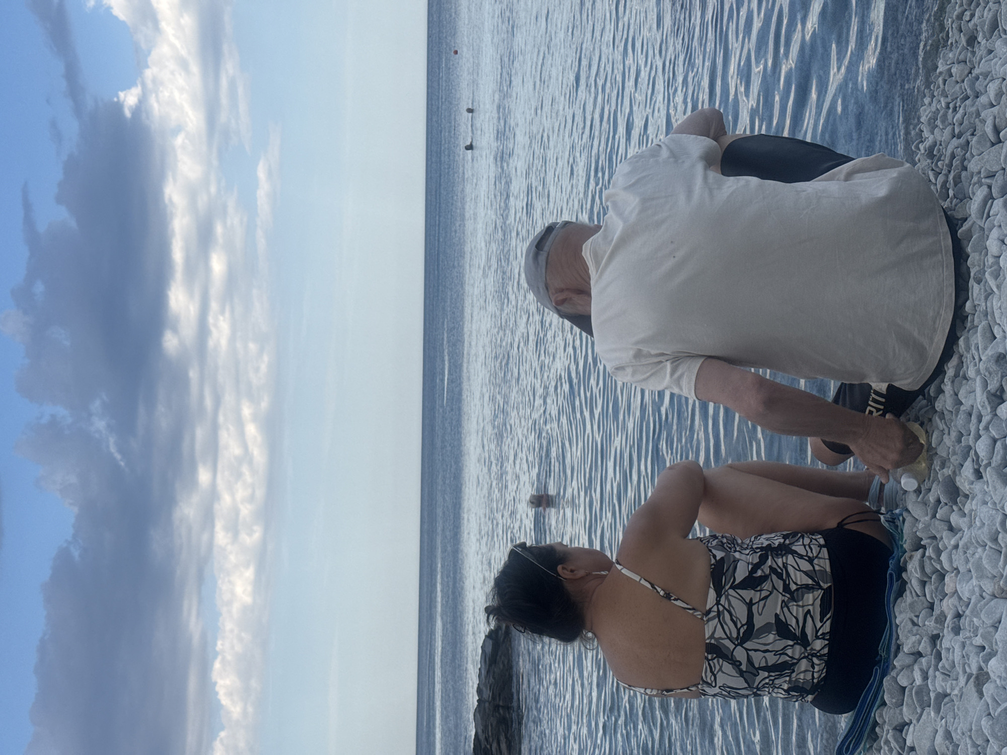In Partenit, Ukraine, on a pebble beach, two people sit facing the calm, rippling sea under a cloudy sky. A woman with dark hair pulled back and sunglasses perched on her head wears a patterned swimsuit and black shorts. Beside her, a man in a white t-shirt and a grey cap sits with his back to the camera. The water is a deep blue, with small waves reflecting the sky. Two figures are visible swimming in the distance, and a red buoy marks the water. The beach is covered in smooth, grey pebbles. A partially opened bottle of liquid sits on the pebbles near the couple. The overall atmosphere is tranquil, suggesting a quiet moment by the sea.