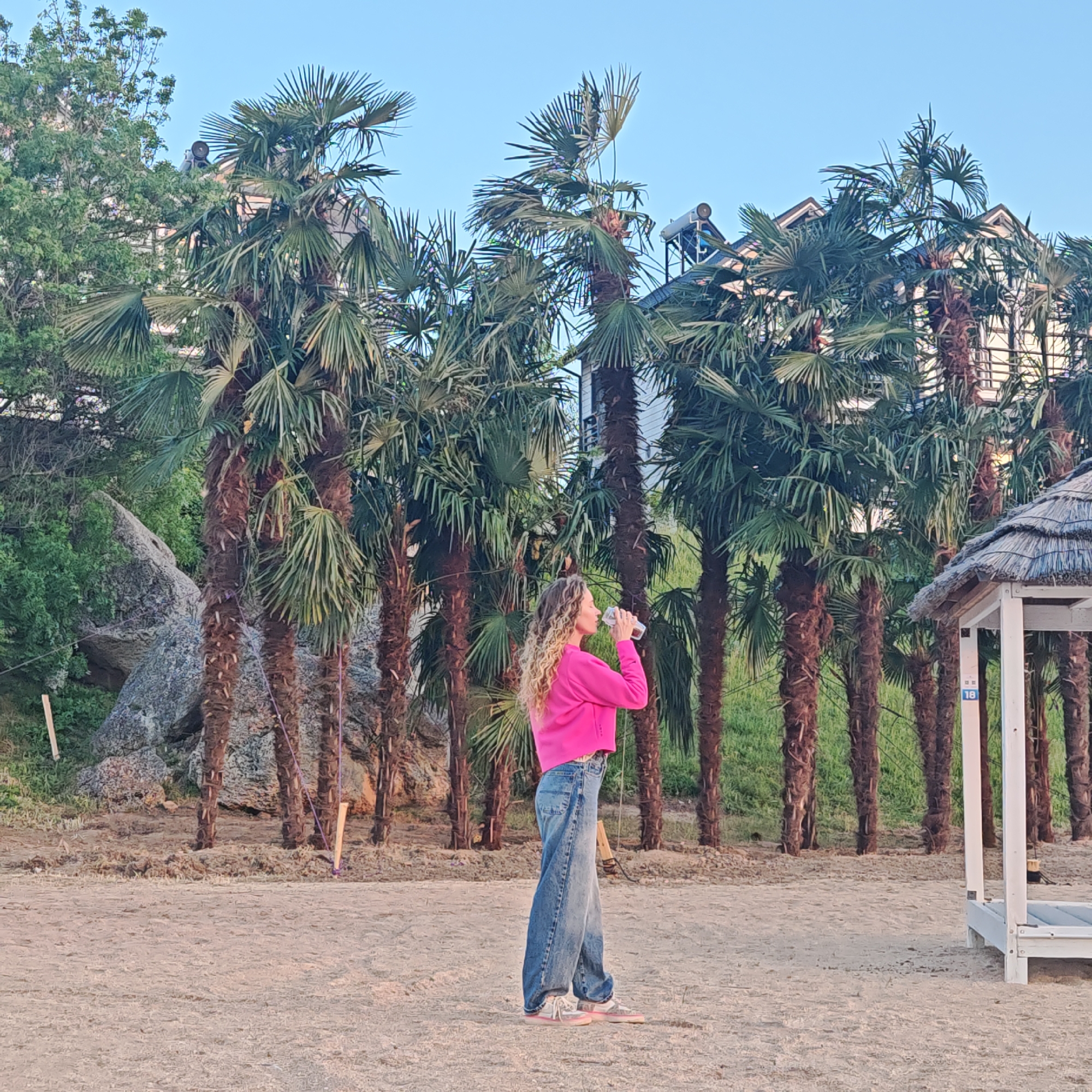 A woman in a pink sweatshirt and blue jeans drinks from a clear bottle on a sandy beach in Мысовское сельское поселение, Ukraine. Palm trees line the background behind her, and a white, partially-visible structure is to her right. A large rock and green vegetation are visible to her left, and building rooftops with solar panels can be seen through the palm trees.