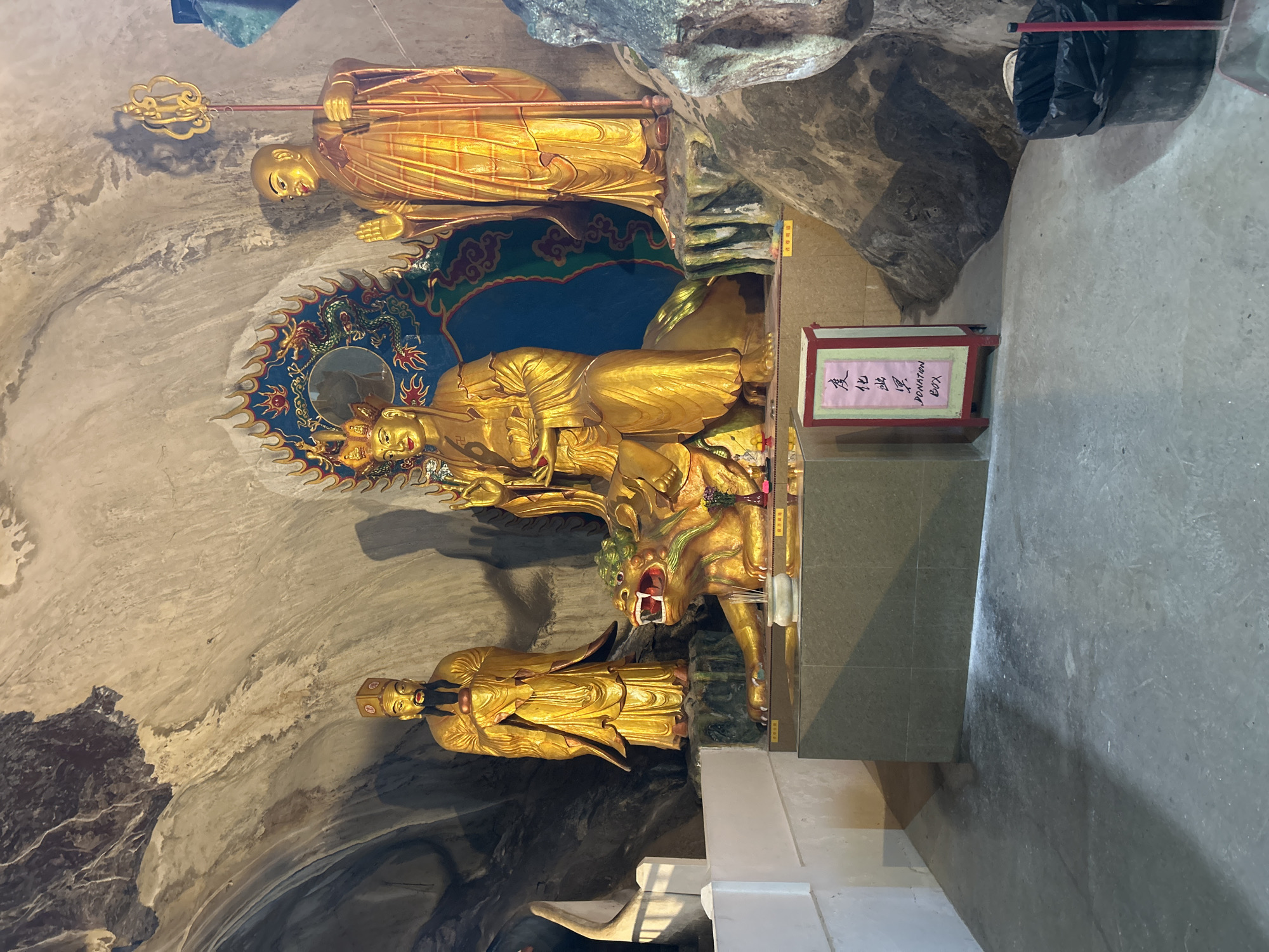The image depicts a cavernous interior, likely a cave temple, with natural rock formations forming the ceiling and walls. The environment appears dimly lit, with artificial lighting casting shadows. In the foreground, the concrete floor is visible, showing some dust and wear.
The central focus of the scene is a shrine area. Three golden statues are prominently displayed on a raised platform. The largest statue, seated and wearing a crown, is positioned in the center. To its right stands another golden statue of a monk-like figure, holding a ceremonial staff. To the left of the central seated figure, a third golden statue depicts a bearded man in traditional robes, holding a round object. In front of the seated figure is a golden statue of a creature resembling a lion or dragon. Incense sticks are burning in a bowl before the shrine.
To the left of the shrine, a white, stepped structure is partially visible. To the right of the shrine, a large, dark rock formation dominates the background. A red and white donation box with Chinese characters and the English text "DONATION BOX" is placed in front of the shrine area. A black trash can with a black plastic liner is also visible to the right of the donation box.
This scene is consistent with the interior of the Sam Poh Tong Cave Temple in Ipoh, Malaysia, which is known for its elaborate cave shrines and statues.