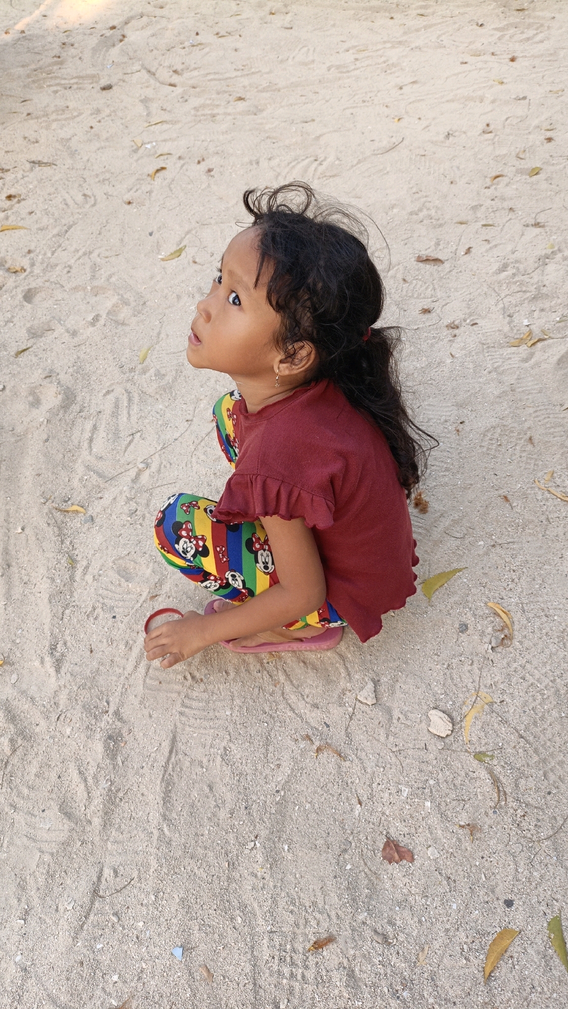 A young girl is captured in a medium shot, squatting on a sandy surface. She has dark, curly hair pulled back into a ponytail and is looking upwards and to her left with wide, curious eyes. She is wearing a maroon t-shirt with ruffled sleeves and brightly colored leggings adorned with a repeating pattern of cartoon characters, including Mickey Mouse. Her feet are bare except for pink flip-flops. The sandy ground is textured with faint footprints and scattered with dried leaves and small pieces of debris. The lighting suggests it is daytime, with shadows indicating the sun is casting light from a high angle. The overall impression is of a casual, outdoor moment, possibly at a beach or park.