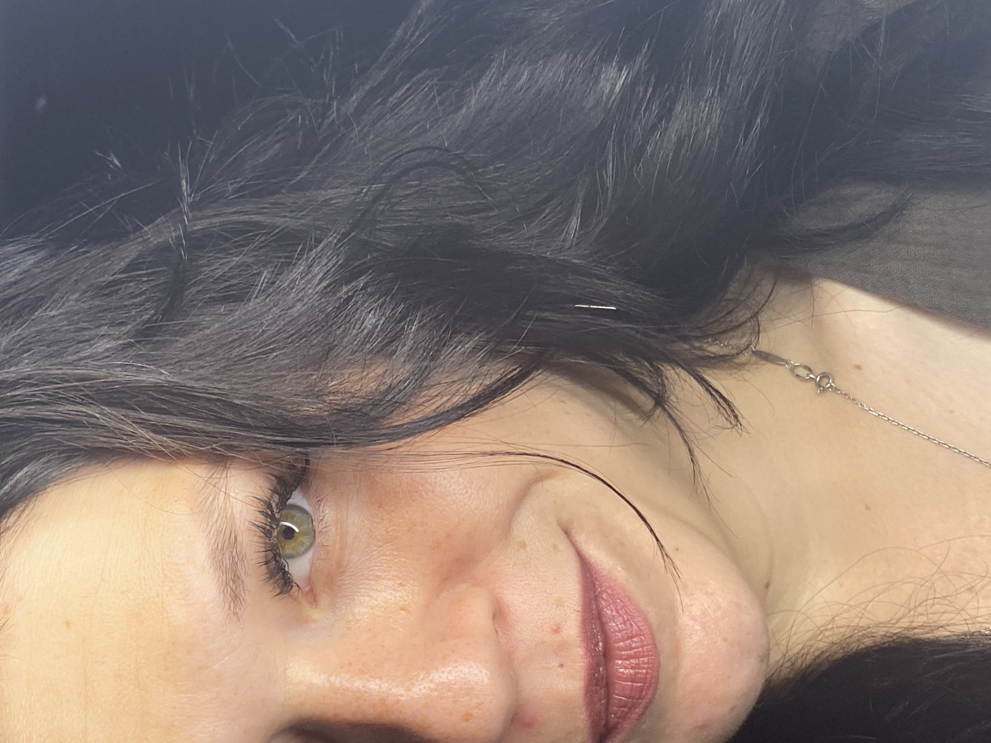 The image is a close-up, portrait-style photograph of a woman's face, captured from the left side of her head. The woman has dark, wavy hair that partially obscures her face. Her skin is fair with some freckles visible on her cheeks and nose. She has green eyes and is wearing makeup, including mascara and lipstick. A thin silver necklace with a pendant is visible around her neck. The background is dark and out of focus, suggesting an indoor setting with low lighting. There is no discernible urban environment or specific location in Chaikovsky, Russia.
