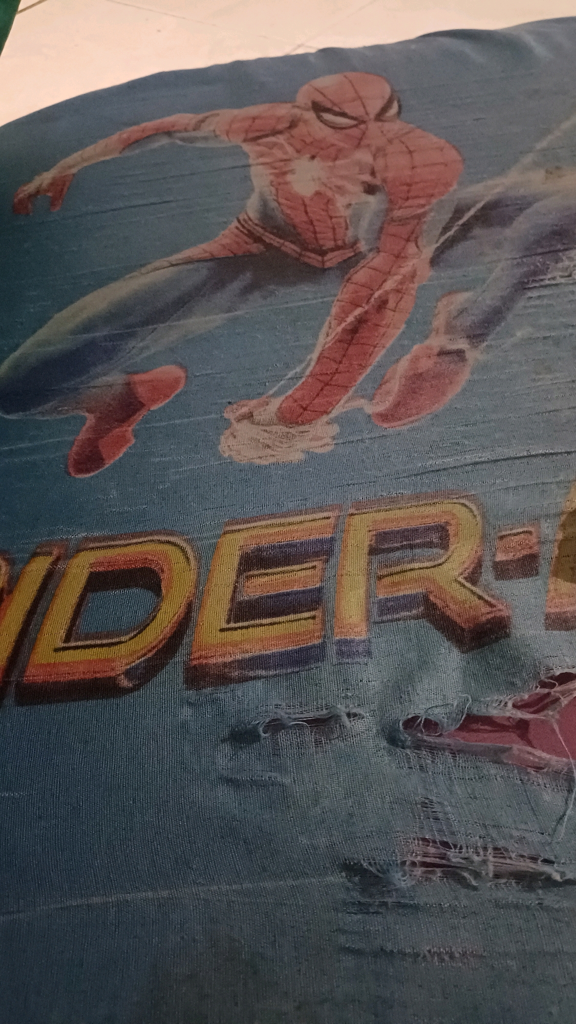 The image displays a close-up of a tattered blue fabric, likely a shirt or pillowcase, featuring a faded graphic of Spider-Man in a dynamic pose. The word "SPIDER" is prominently displayed in large, colorful, gradient letters below the character. The fabric exhibits significant wear and tear, with multiple holes and frayed edges, particularly in the lower right quadrant. The lighting suggests an indoor setting, possibly a bedroom or laundry area, with natural light falling from the top left. There is no indication of a specific location within the city of Wonokerso, Indonesia.