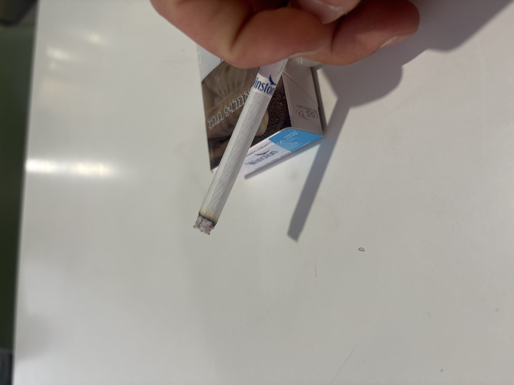 An overhead shot captures a hand holding a lit cigarette and its open pack on a smooth, reflective light-colored surface, likely a table or desk, in an indoor setting. The hand, with light skin tone, holds the cigarette towards the lower left of the frame, with the cigarette pack positioned above it.
The cigarette, primarily white with a blue "Winston" logo, is partially smoked, showing a glowing, reddish ash at its tip. The cigarette pack, also branded "Winston," features blue and brown accents and is partially open, revealing more cigarettes inside. Several pieces of text are visible on the pack: the "Winston" brand name is prominent, a blue tab reads "FRESH SWIPE," and small letters/numbers like "CE SS" are also present. Additionally, there is text in a non-Latin script, specifically Hebrew, which reads "העישון מזיק לבריאות" (Smoking is harmful to health).
The overall activity depicted is a person holding a lit cigarette and its pack, suggesting a moment either before, during, or after smoking. The scene is clean and simple, with no specific time of day or weather discernible from the indoor environment. The image is provided with the location context of Yerevan, Armenia.