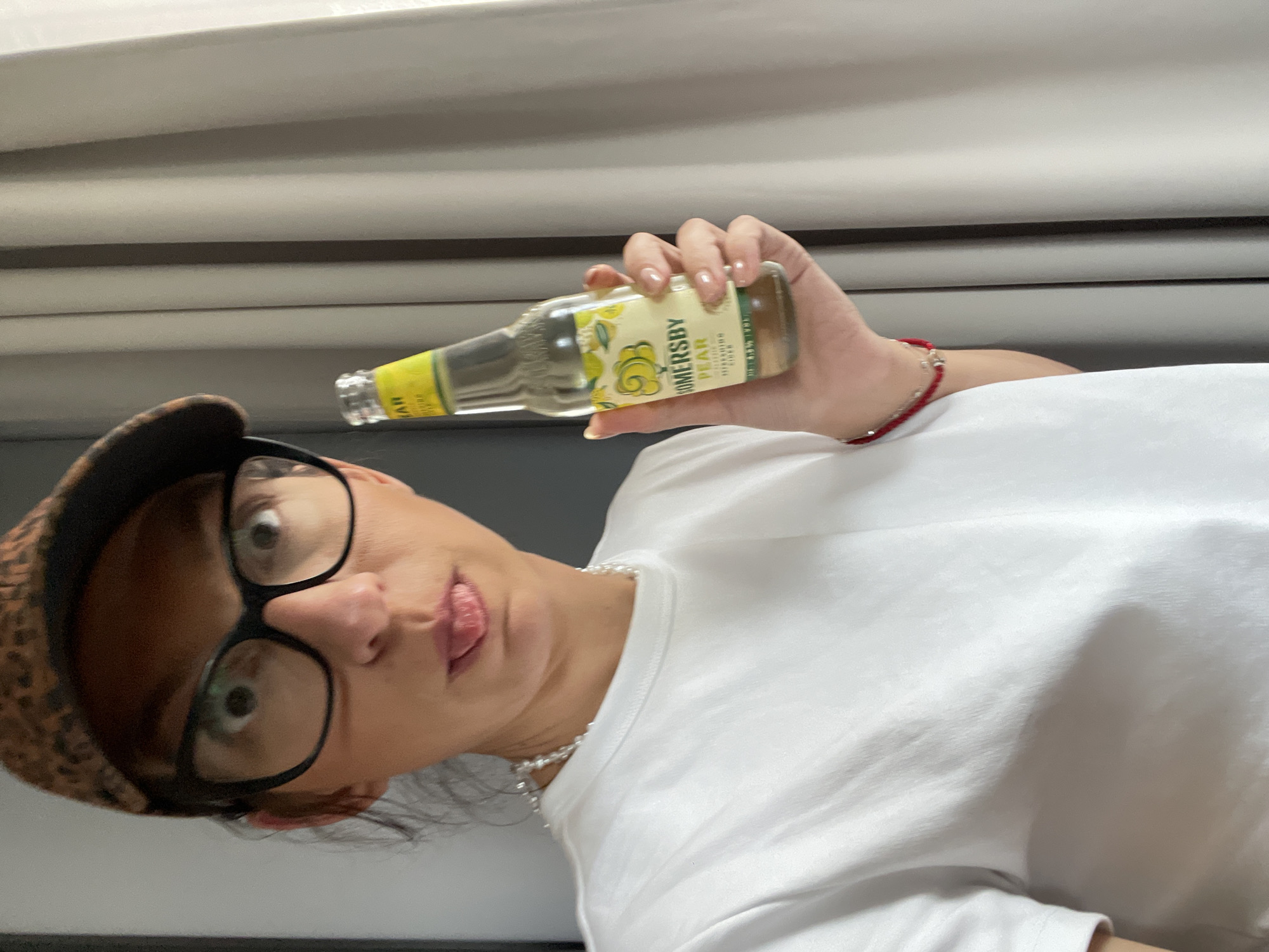 A woman is holding a bottle of Somersby Pear hard cider. The woman has fair skin, dark hair and eyes, and wears a white t-shirt, a leopard print hat, and large-framed glasses. She is wearing a red bracelet and a pearl necklace. She is making a silly face, sticking out her tongue slightly. The background is a light grey blind. The image was not necessarily taken in Sofia, Bulgaria.