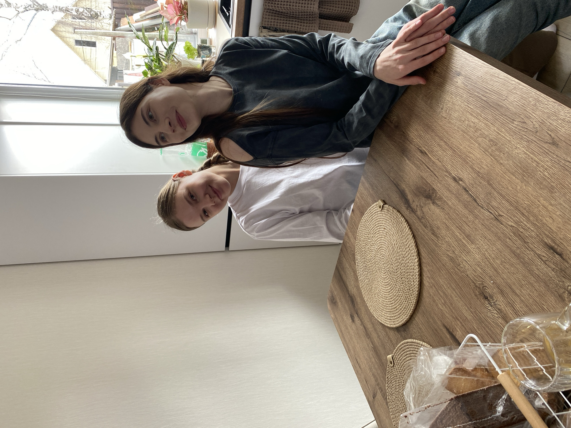 Two young women sit at a wooden table. One woman is wearing a long-sleeved gray shirt and jeans. The other is wearing a white shirt. On the table are two woven placemats and a bread rack containing bread. A window shows a view of what appears to be a residential area. The location is Votkinsk, Russia.