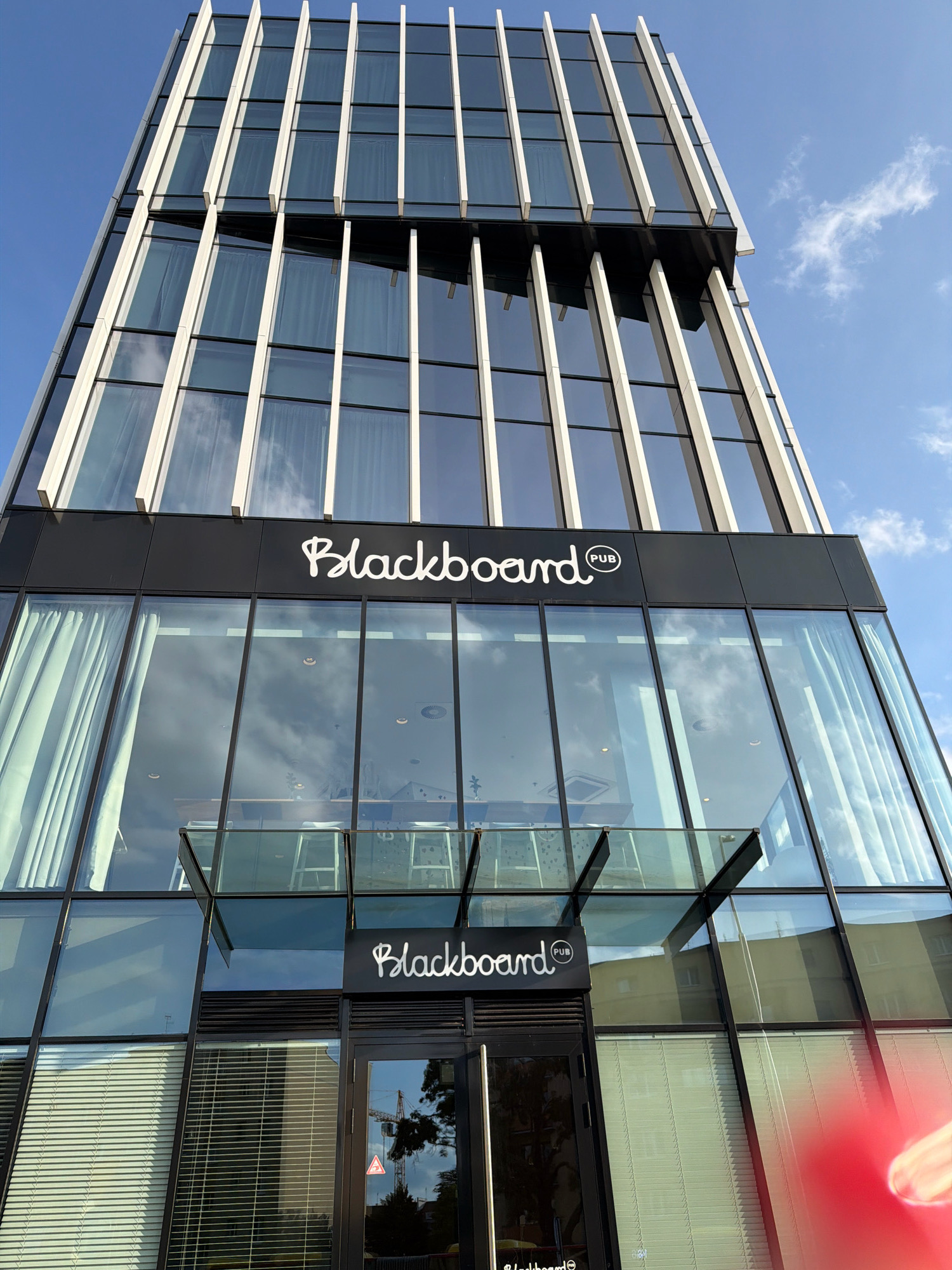 The image shows the exterior of the Blackboard Pub, a business located in Wrocław, Poland. The pub is situated in a modern glass-fronted building. The facade features large glass windows, reflecting the sky and clouds. A prominent sign with the pub's name, "Blackboard PUB," is visible on the upper part of the building. The lower part of the building also has a similar sign above the entrance. The building has a contemporary architectural style with vertical white elements accentuating the glass facade. The sky is clear with a few scattered clouds. The overall scene depicts a commercial establishment in an urban environment.