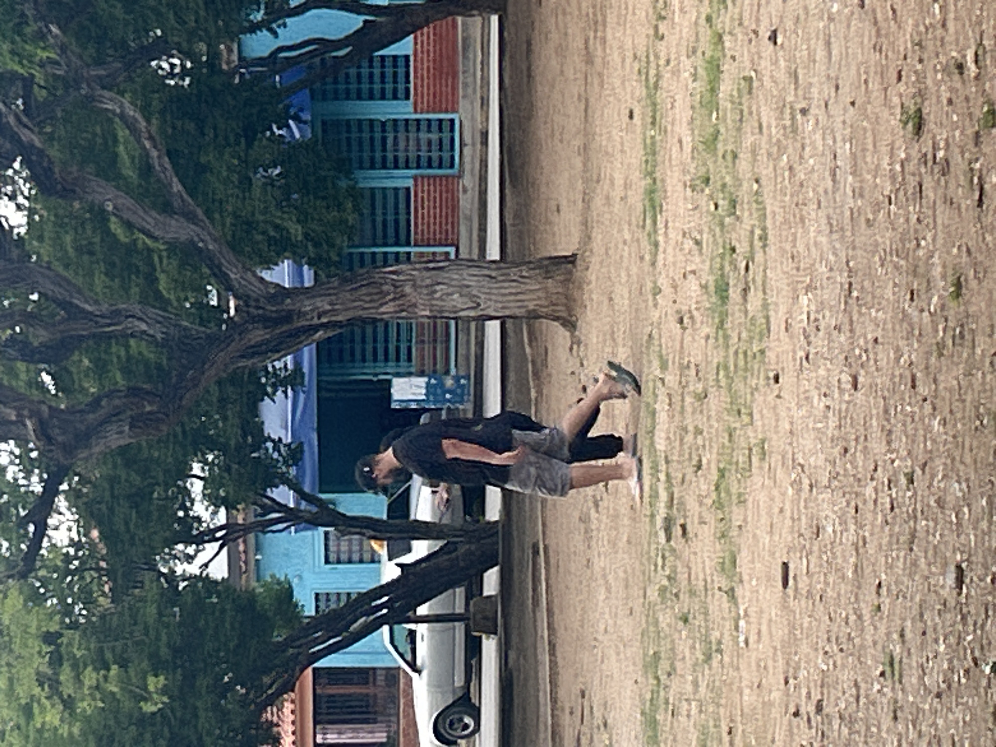 The image captures a street scene in Santa Ana, Venezuela. Two individuals are walking from left to right across a dirt and sparse grass area. The person in front, a man wearing a black t-shirt, gray shorts, a black baseball cap, and flip-flops, is looking towards his right and holding a mobile phone. He is accompanied by a second person, whose lower body is visible from the waist down, wearing dark pants and sandals. Behind them, a white car is parked on the street, and further in the background, a light blue building with red brick accents and striped windows is visible. Large trees with textured bark and dense green foliage frame the scene, with their branches extending across the top portion of the image. The ground is covered in dry dirt with patches of green grass and scattered small stones and debris.