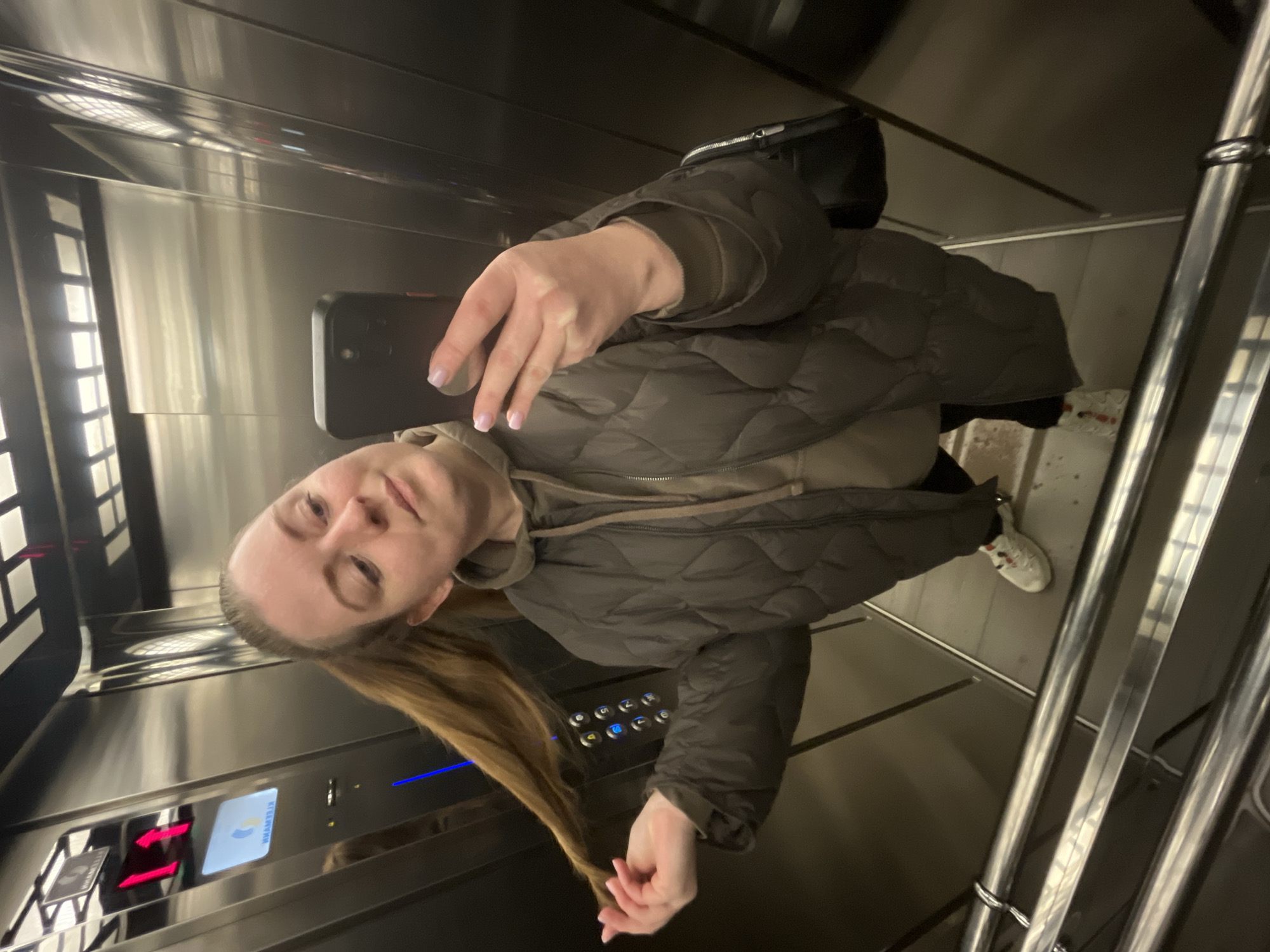 An adult female is depicted inside a modern elevator located in Klin, Russia, taking a selfie. She has long, light brown hair styled in a high ponytail, with her left hand holding a section of her ponytail. She wears a dark olive green quilted puffer jacket over a lighter brown hooded sweatshirt, dark trousers, and white sneakers with red and black details. Her right hand holds a black smartphone in a black case, angled towards herself to capture the image. Her fingernails are painted a light pink shade. A small black shoulder bag with a silver zipper is visible on her right side.
The elevator interior features highly reflective, dark metallic walls and a ceiling with integrated grid-patterned lighting. On the left wall, a control panel is visible. It includes a red digital display showing "1" with an upward arrow, indicating the elevator's floor or direction. Below this, a screen displays a mirrored "KLEEMANN" logo. A set of circular buttons with blue backlighting is also present. The floor is dark, and two polished metal horizontal handrails are installed in the lower portion of the cabin.