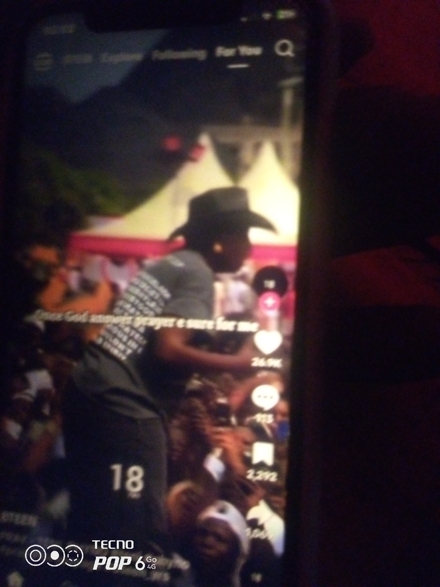 The image shows a close-up of a smartphone screen displaying a video. The video features a person wearing a dark cowboy hat and a grey t-shirt with a white pattern. The person is in the foreground, appearing to be in a crowded outdoor setting with tents in the background. Text overlays on the video include "For You", "26.9K", "913", "2,292", "1,004", and "18". At the bottom left corner of the phone screen, the TECNO POP 6 4G logo is visible. The overall scene appears to be a public gathering or event. The location in the city of Howayi, Nigeria is not depicted or inferable from the provided image.