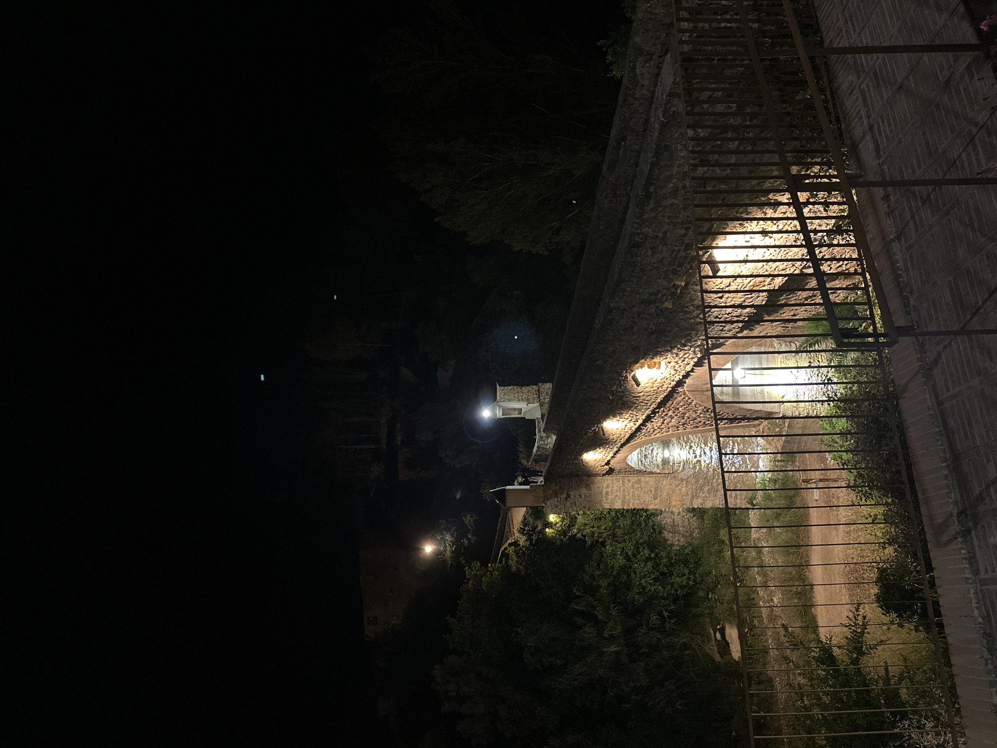 The image shows a night view in Montanejos, Spain. A stone aqueduct is illuminated by lights that shine from underneath the stone arches. A metal fence runs along the aqueduct on the right. Trees and foliage are visible in the background. The foreground is dark, with some illuminated structures visible in the distance. The aqueduct is the primary subject, with the fence, lights, and surrounding vegetation contributing to the overall scene.