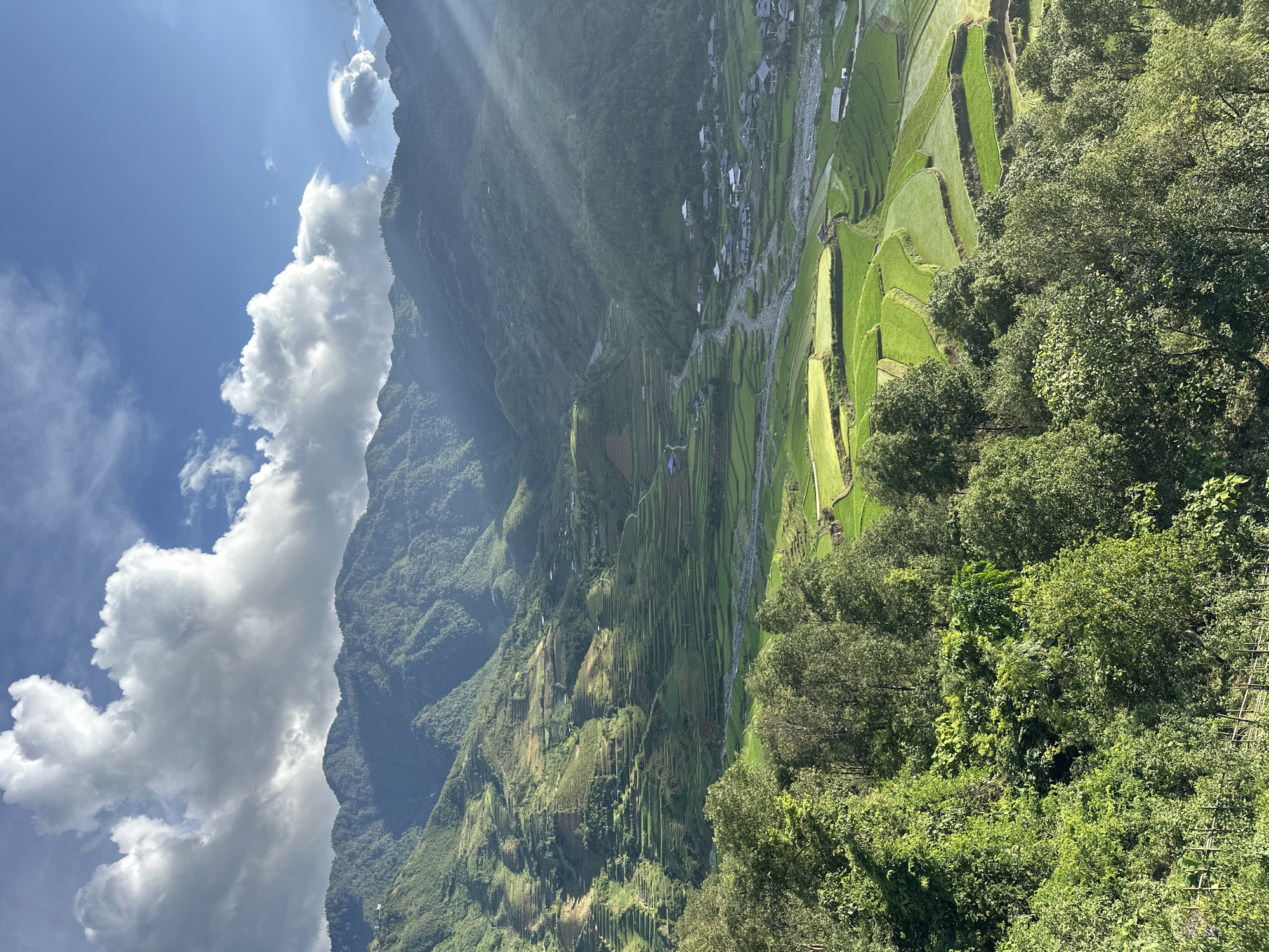 A high-angle, wide shot captures a verdant mountainous valley in Tú Lệ, Vietnam, under a partly cloudy sky. The upper portion of the frame features a bright blue sky partially covered by large, white and grey cumulus clouds. Sun rays are visibly descending from the upper right, piercing through the atmospheric haze. The background is dominated by multiple layers of densely forested, dark green mountains, their slopes displaying some signs of agricultural terracing at higher elevations.
The midground reveals an extensive network of bright green rice terraces that conform to the natural contours of the valley walls and floor. A light-colored, rocky river or stream meanders through the center of the valley, separating sections of the terraced fields. Adjacent to the river, a compact village cluster is visible, composed of numerous houses with dark-colored roofs. Narrow paths or roads are discernible winding through the terraced landscape and connecting the village structures. The foreground is filled with a dense canopy of lush, dark green trees and foliage, indicating an elevated viewpoint overlooking the valley. No human figures or discernible activities are present in the scene.
