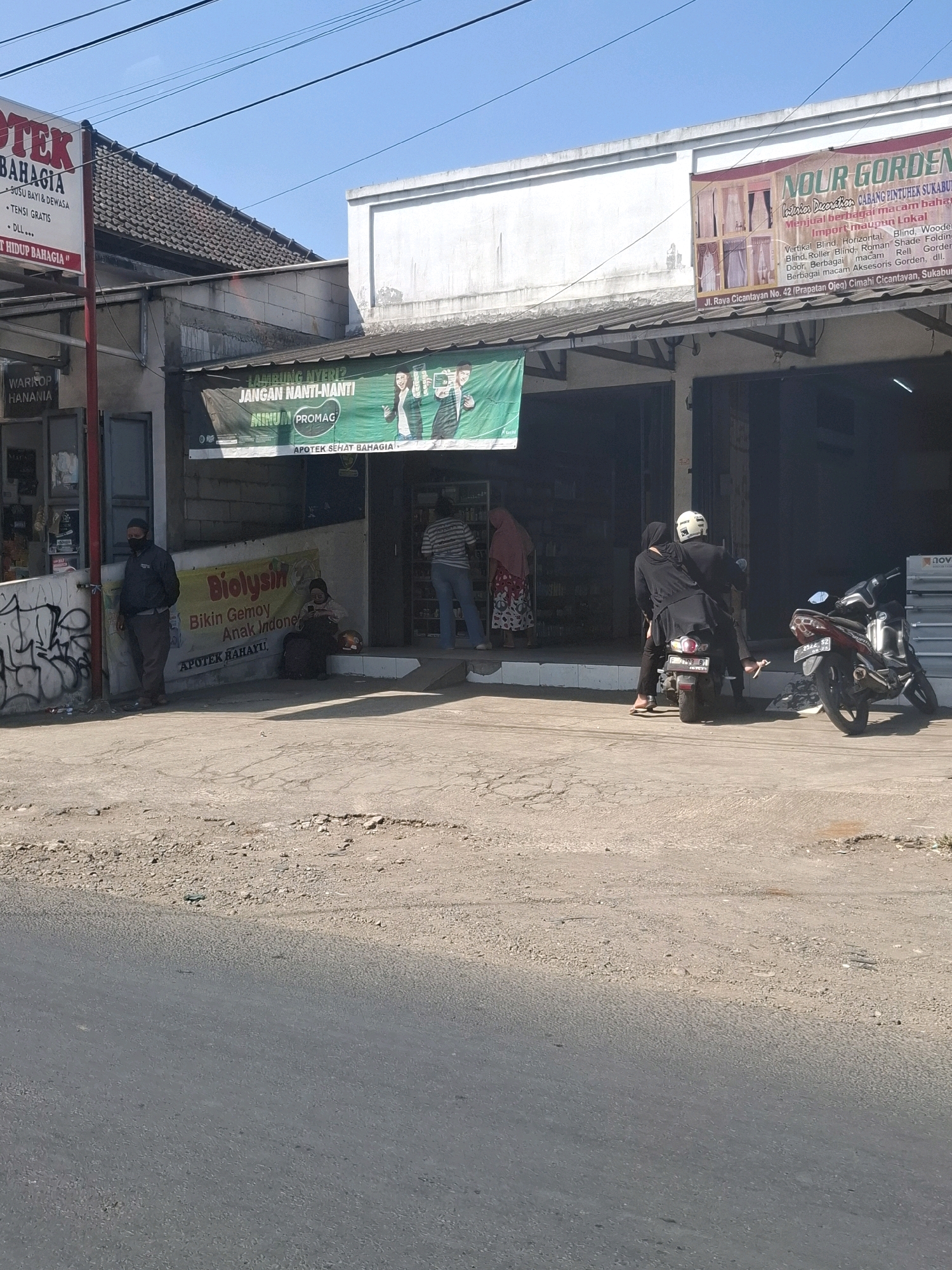 The image depicts a street scene in Kutasirna, Indonesia, on a clear day. In the foreground, a cracked asphalt road leads towards a row of shops. Two motorcycles are parked on the side of the road. One motorcycle, dark red and black, is parked on the right. Another motorcycle, black, is further down the street, with two individuals on it, one wearing a white helmet.
The shops in the background have awnings. One shop, "NOUR GORDEN," displays curtains. Another shop has a green banner advertising "JANGAN NANTI-NANTI MINUM PROMAG" and "APOTEK SEHAT BAHAGIA." To the left, a sign for "OTEK BAHAGIA" and "WARUNG HANANIA" is visible. A man wearing a dark jacket and a face mask stands near the entrance of one of the shops. Inside, shelves of products are partially visible. A woman is seated on the ground in front of a shop, looking at her phone, with a helmet next to her. Another person in a pink and white patterned dress stands near the entrance of the shop with the green banner. The environment appears to be a commercial area with moderate pedestrian and vehicle activity.